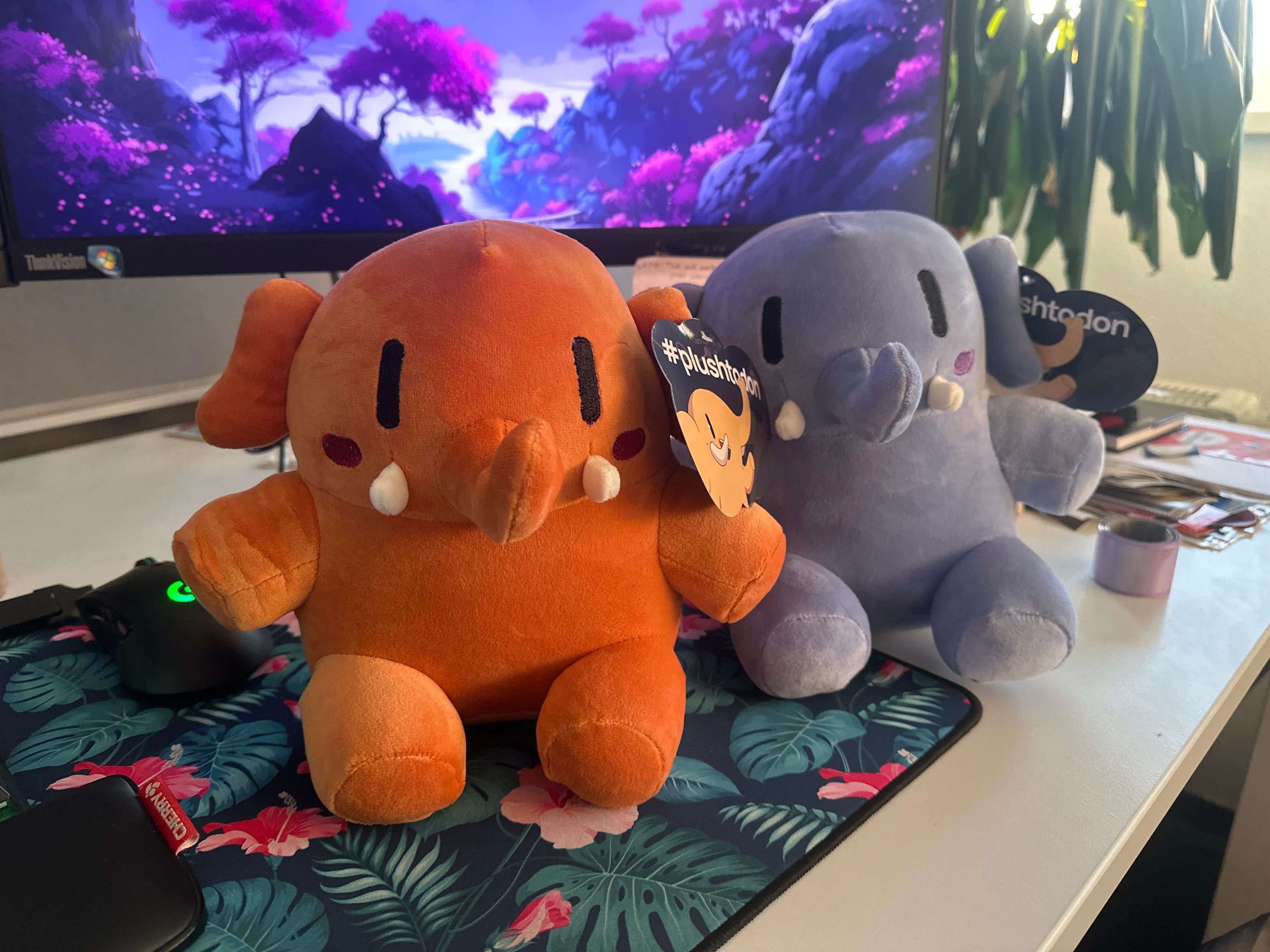 Two plush mastodon toys sit on a desk, one orange and one purple, both featuring tags that read "#plushtodon." A computer monitor with a colorful landscape background is visible behind them, along with a gaming mouse and a floral-patterned mousepad.