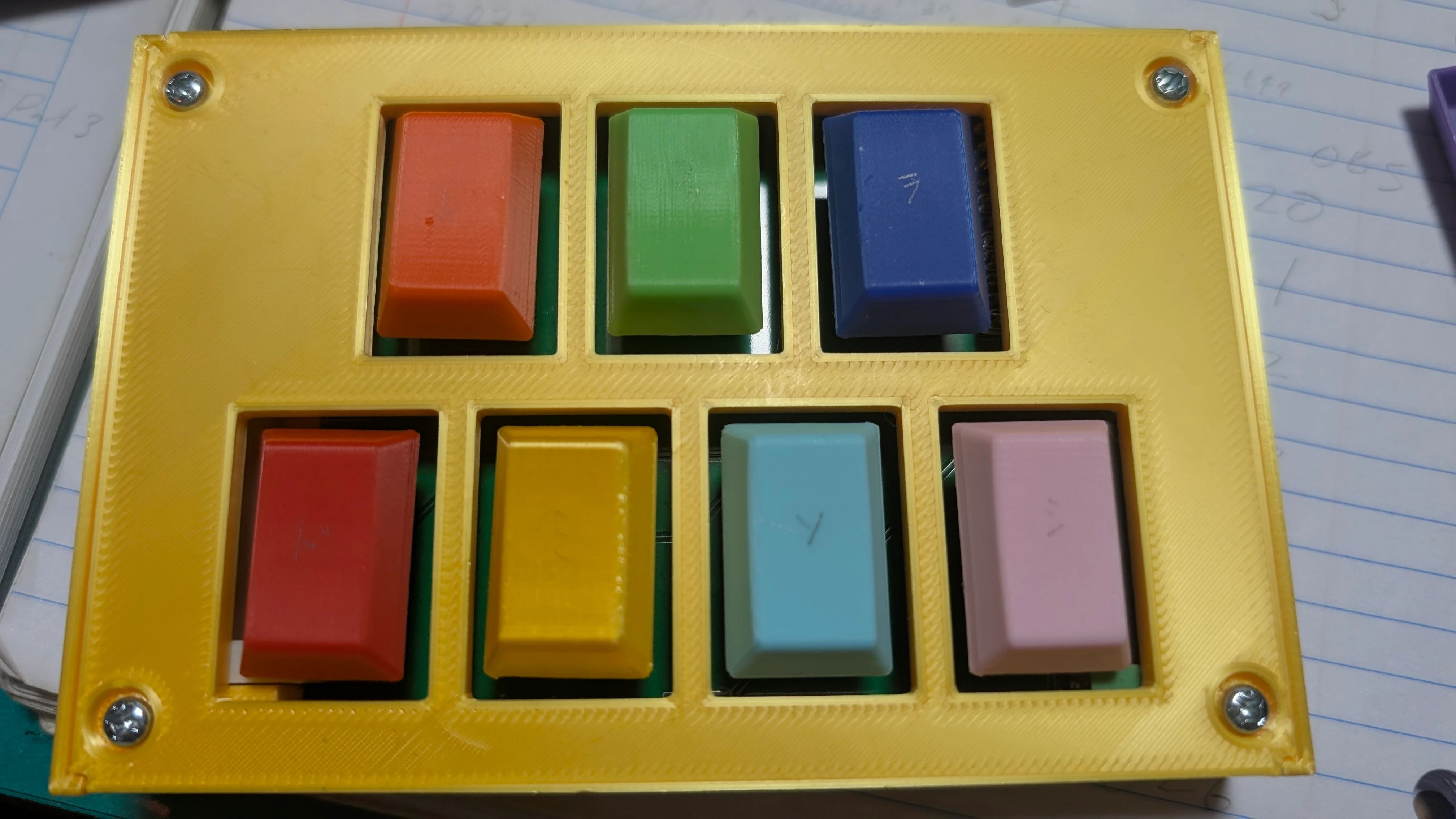 A yellow 3D-printed plate with four screws at the corners holds seven rectangular keycaps in cutouts arranged in two columns. Left column, top to bottom: red, yellow, light blue, pink. Right column: orange, green, dark blue.