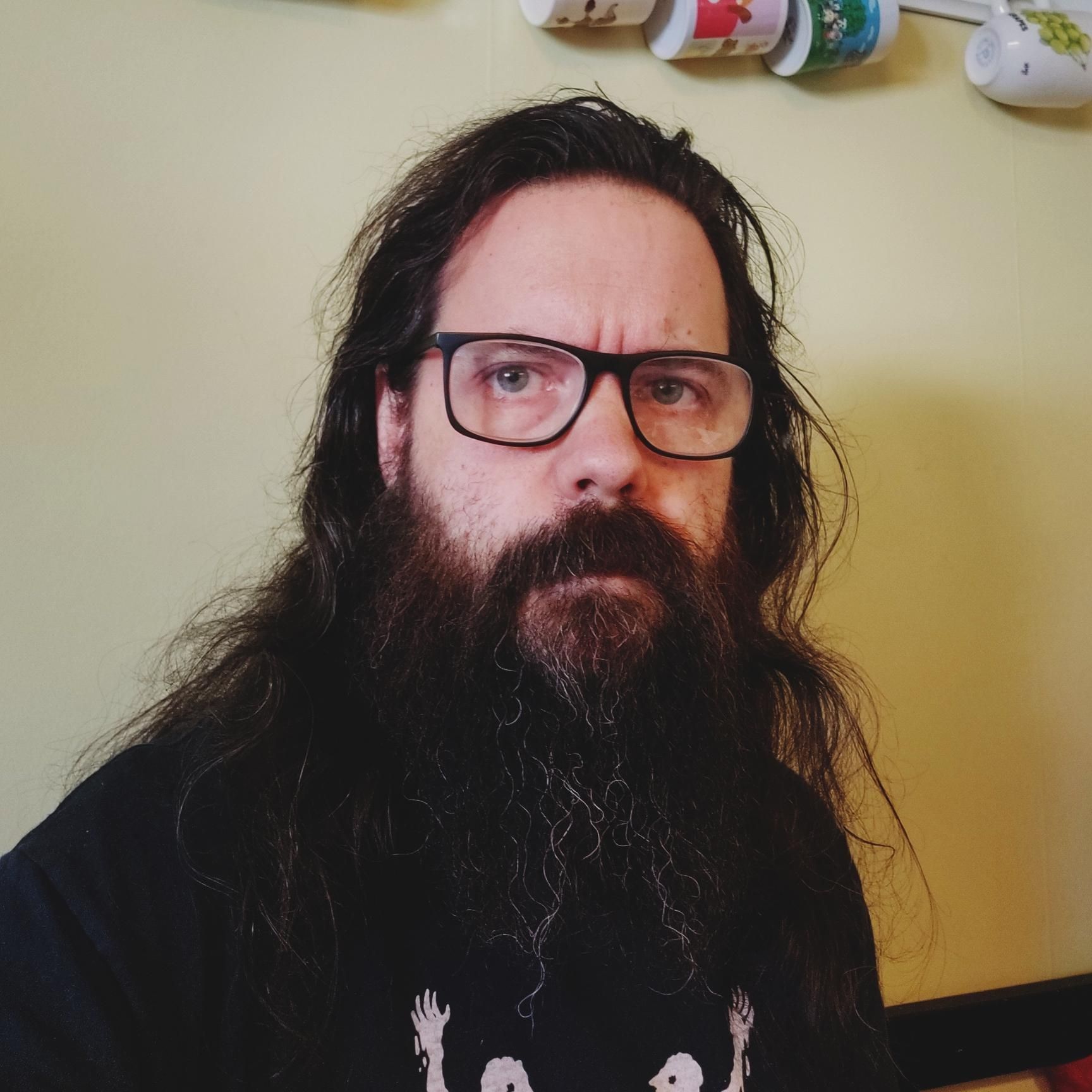 A photo of a surly middle aged man with long dark hair and beard wearing glasses. 