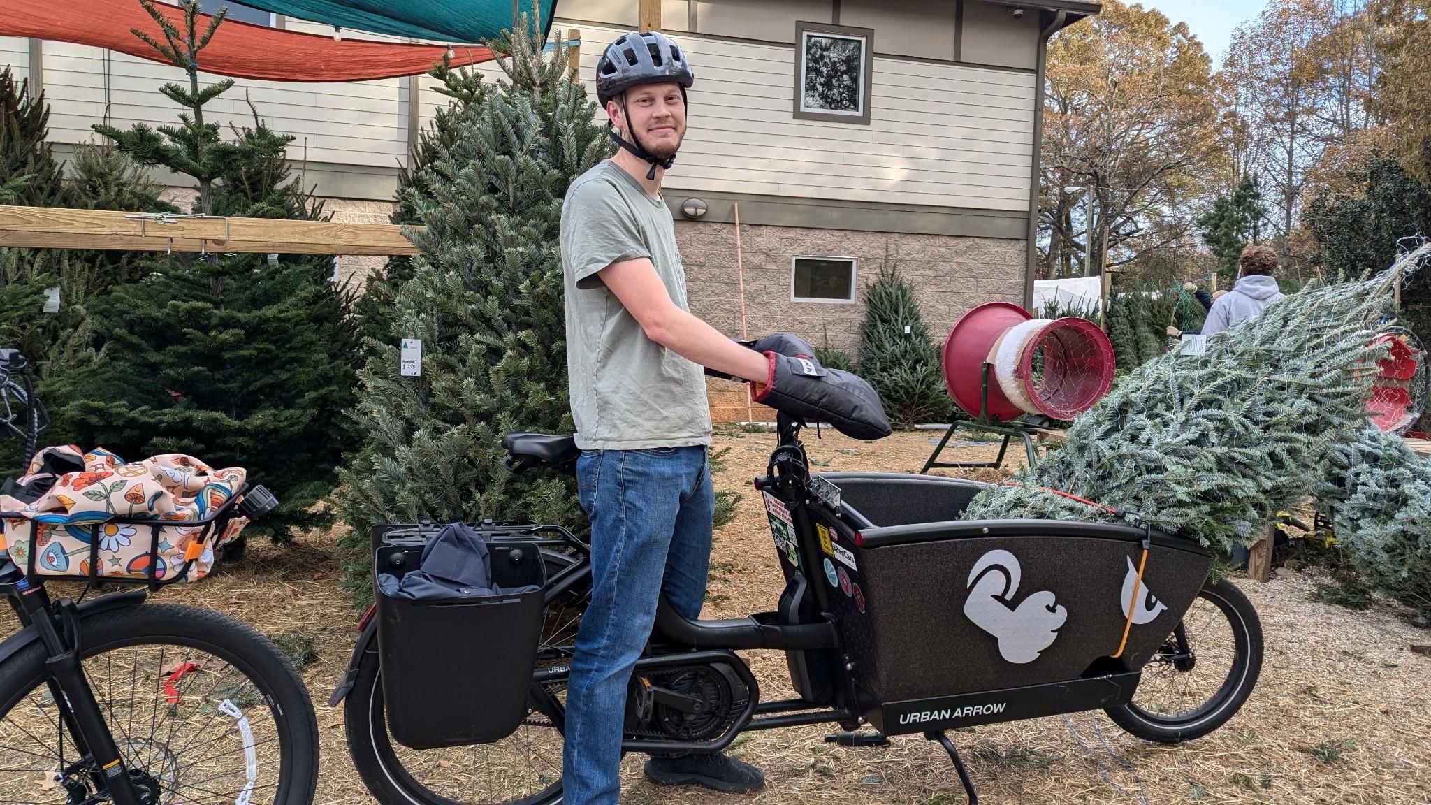 Man (me) standing with a cargo bike with a Christmas tree inside the front bucket. Christmas trees in background. 