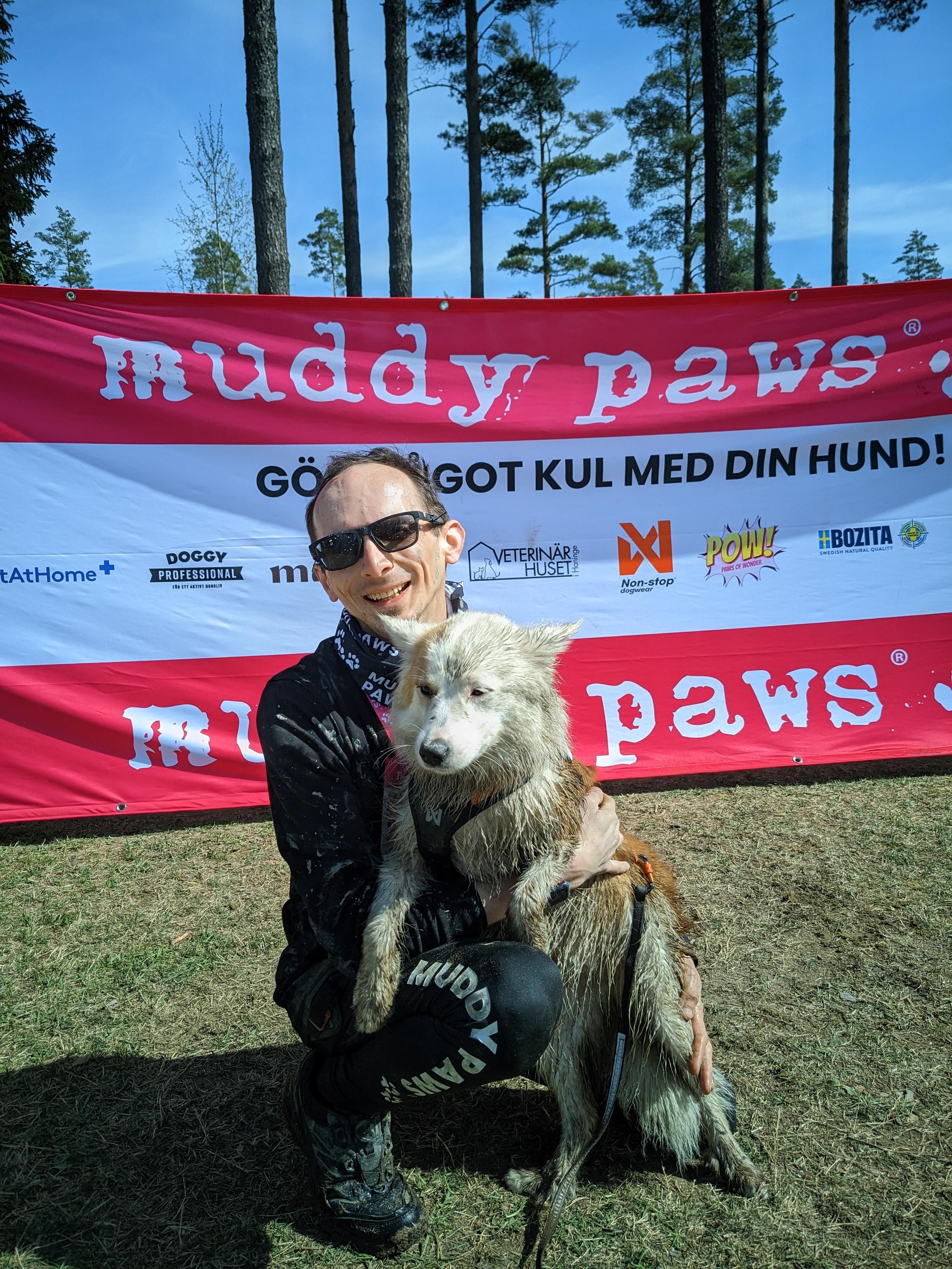 Extremely muddy dude in sunglasses, his face with a grin from ear to ear. He is kneeling with her small (originally) white & red, mud-covered pup in front of a big sign that reads "Muddy Paws"