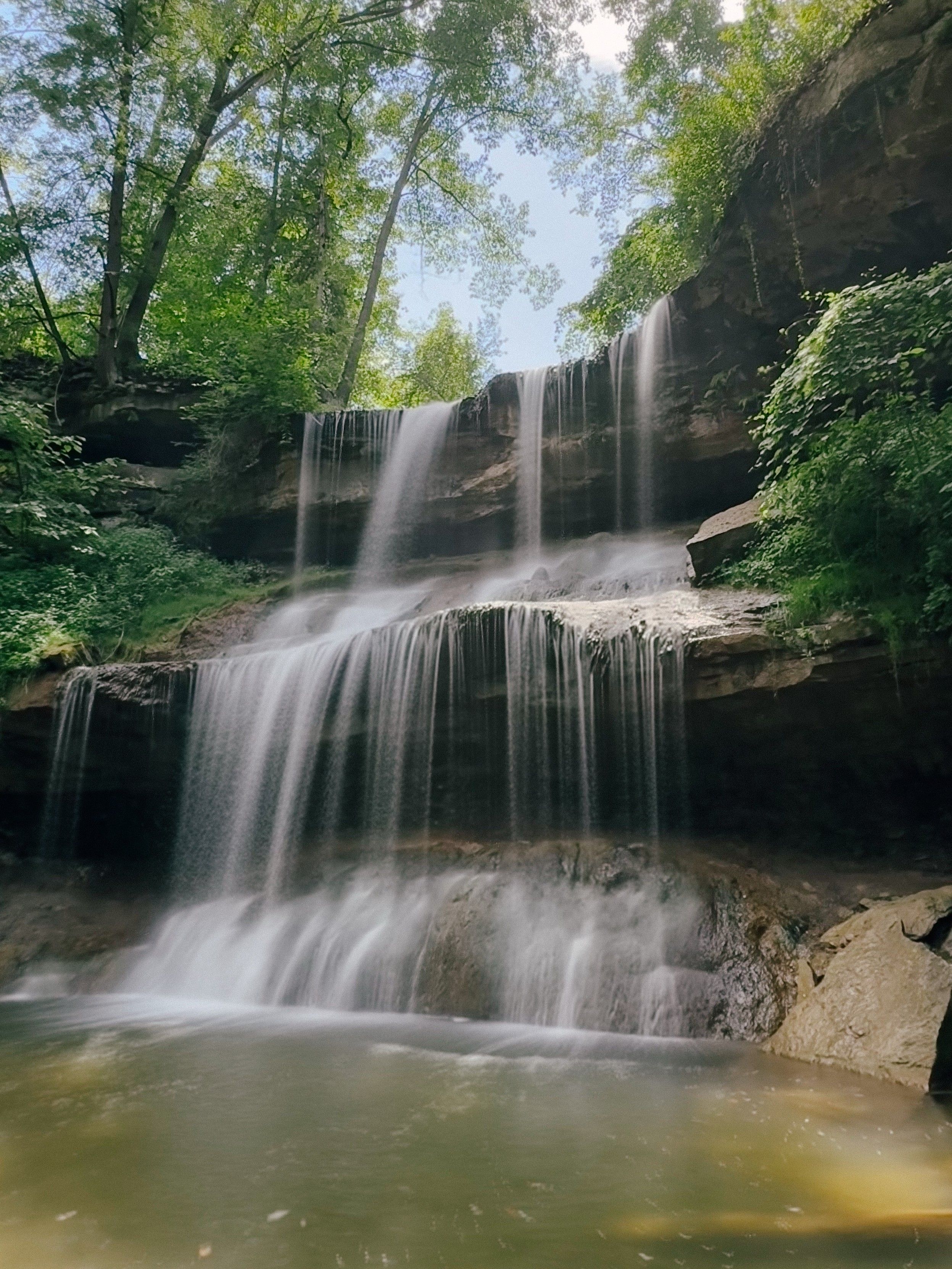 The same waterfall from the other side. There are two strata of rocks in a horseshoe shape the water is flowing down. Lots of plants are growing on the second stratum and trees are on the top level. There is a large pool at the bottom great for swimming. A large tree is submerged on the bottom right creating a great spot for fish to congregate. 
