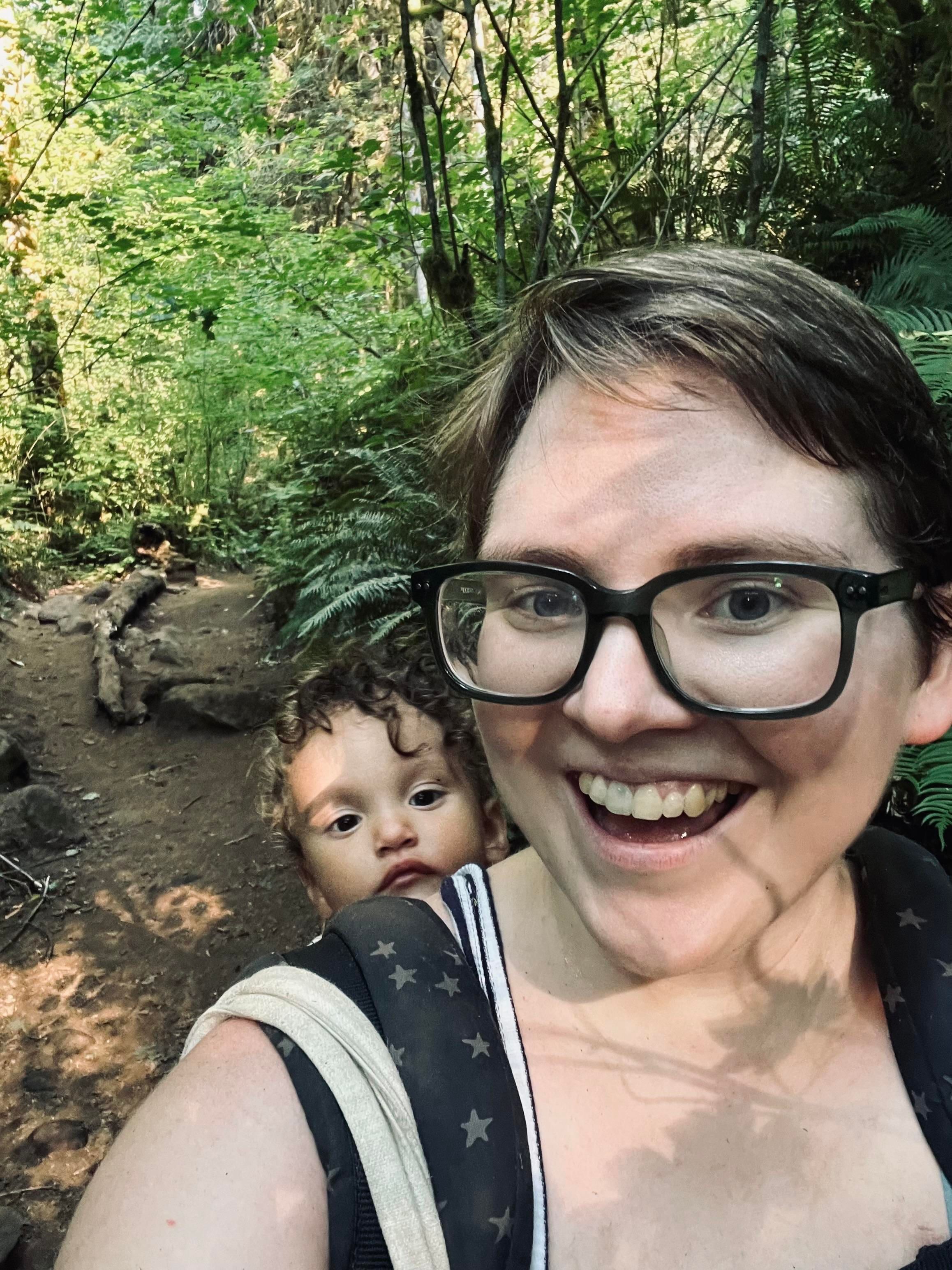 A white person with short hair and glasses smiles at the camera while carrying a child in a back carrier. They are surrounded by lush green foliage in a forested area, with a dirt path visible in the background.