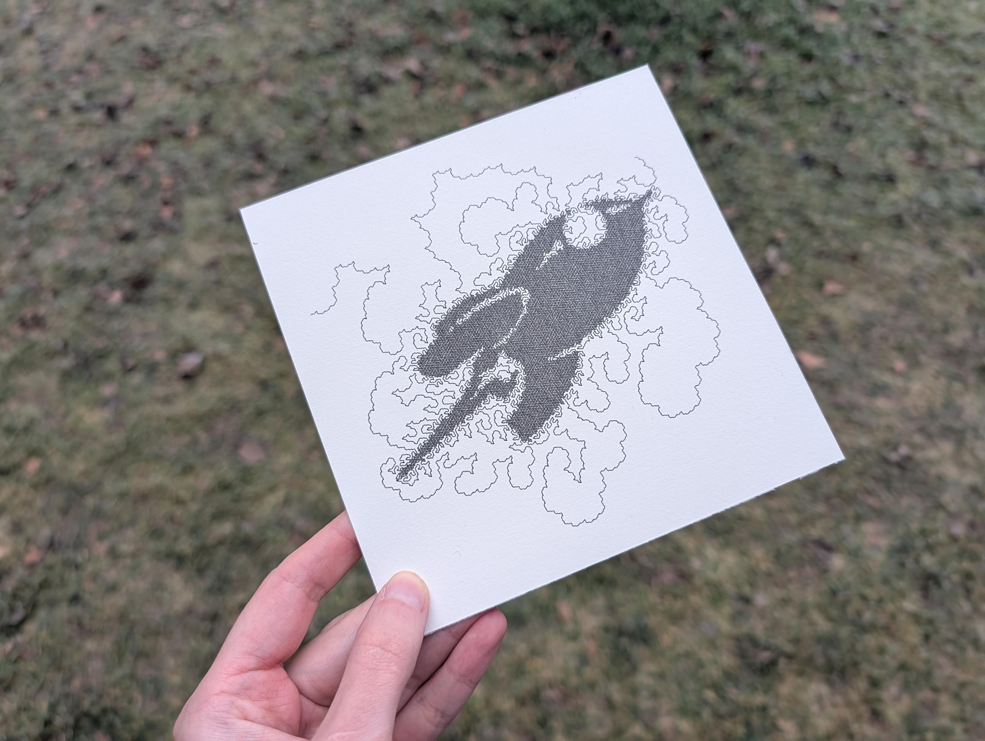 A white hand is holding a square piece of paper – it shows a space-filling curve that looks a bit like the outlines of clouds. It gets more dense towards the center, and that way, clearly shows the shape of Fairy Dust, the mascot rocket of the CCC.