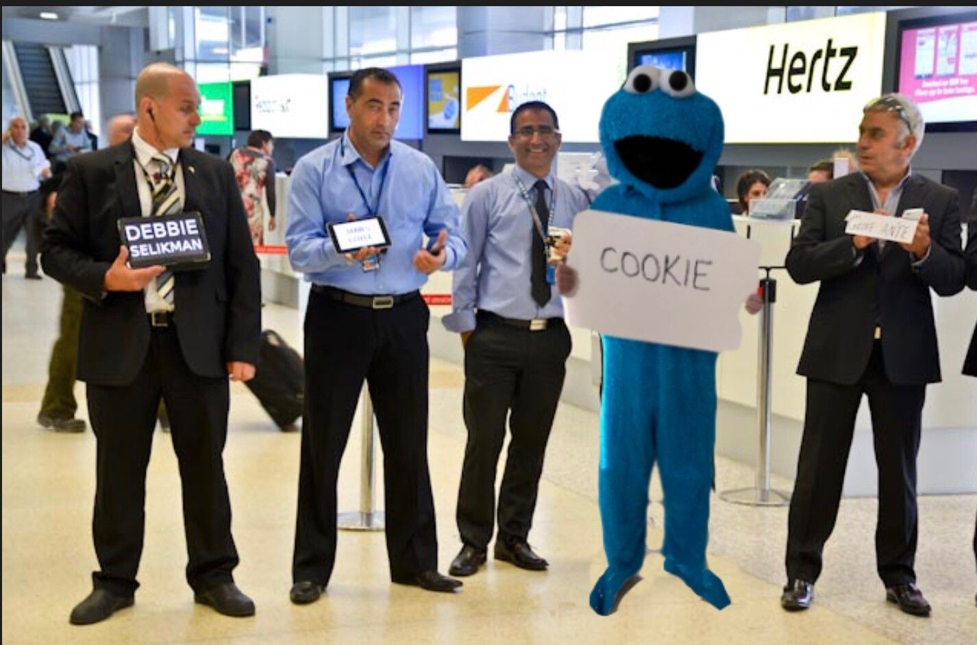 5 people, 4 men in suits and one dressed as Cookie Monster, standing in an airport terminal holding up signs with passenger names on them.
Cookie Monster's sign simply says COOKIE