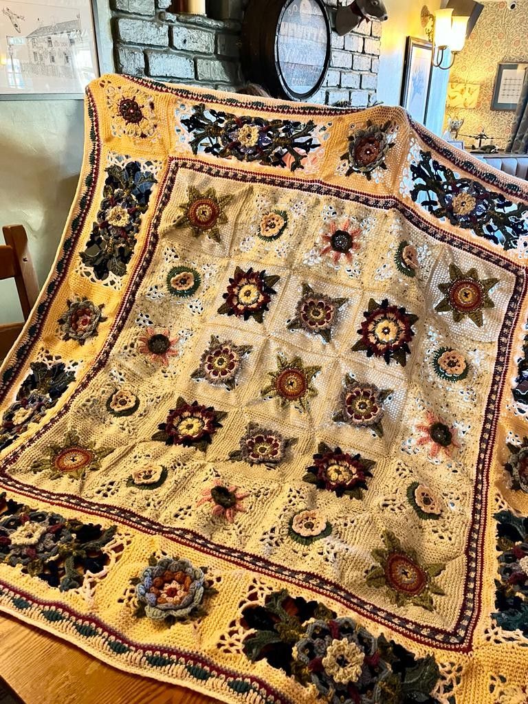 Crochet blanket being held up towards the camera, in an English pub bar. 
The blanket has various crochet flower motifs 