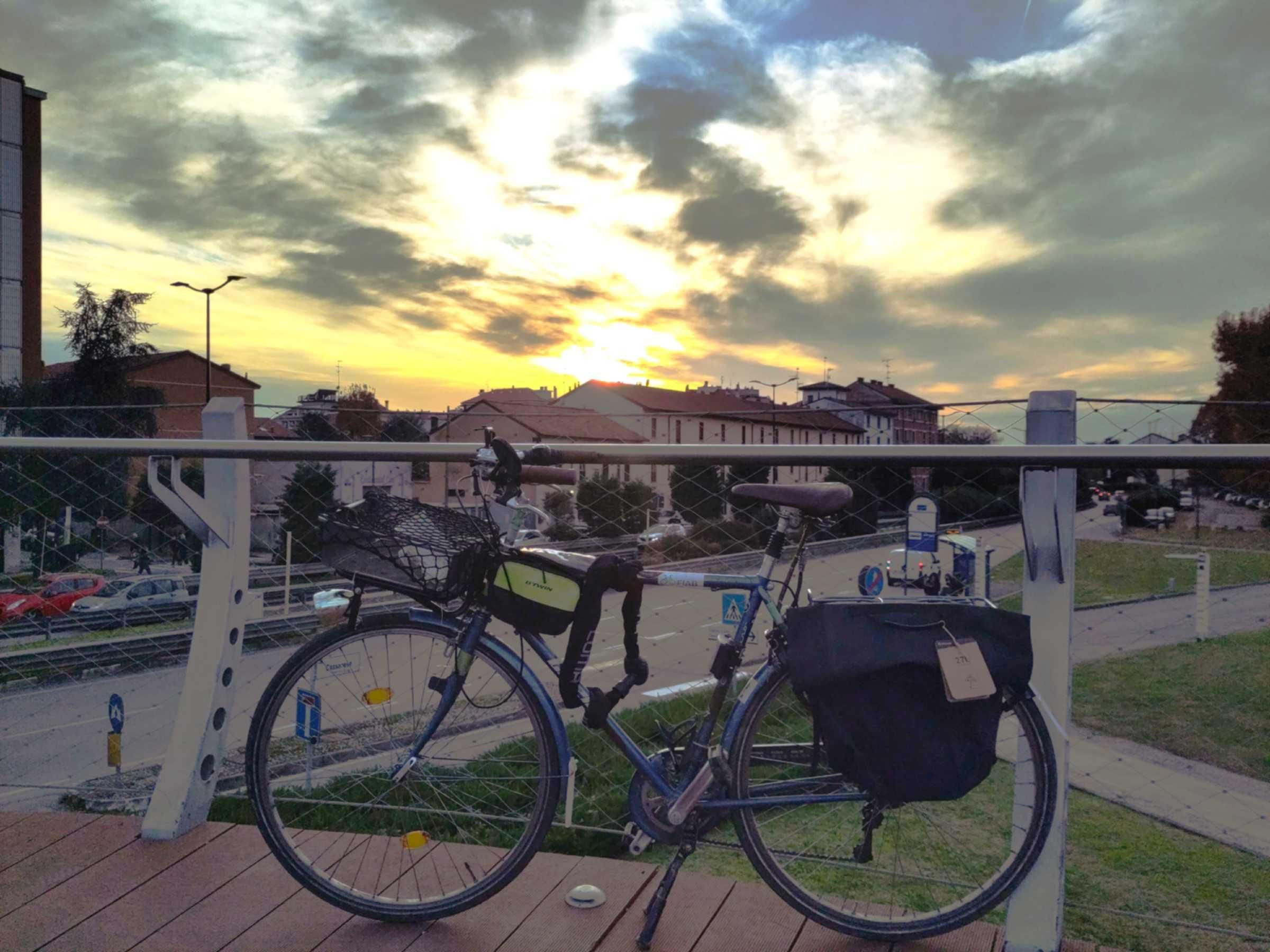 Foto della mia bici blu incorniciata sopra una passerella ciclopedonale. Dietro si vede una strade e alcune case. Sopra il cielo è nuvoloso, ma l'aria è limpida e i colori accesi. In basso nel cielo domina il giallo del sole, in alto l'azzurro