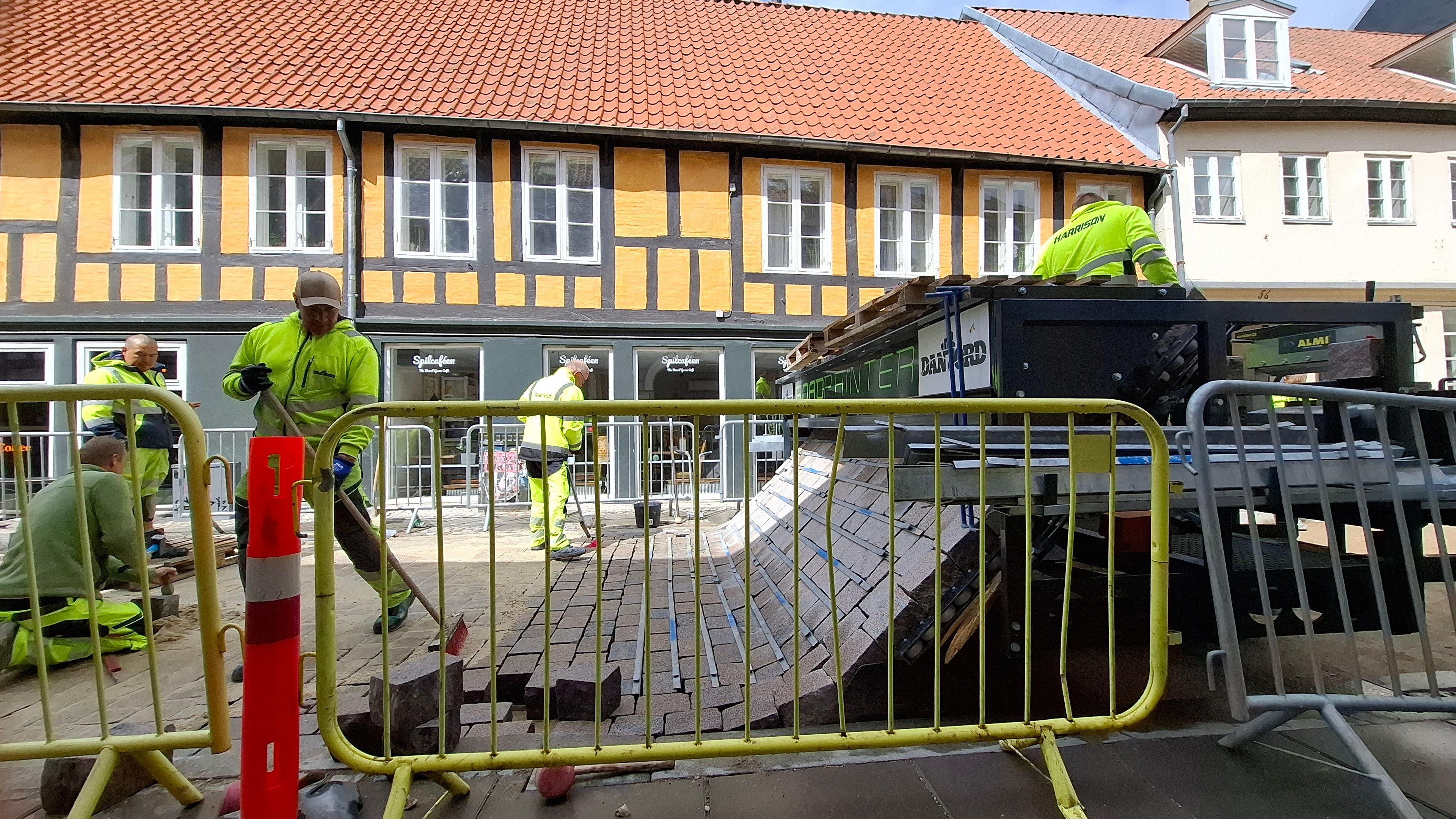 Vestergade, mid-city Aarhus, Denmark. A lovely narrow old street that is, after a few years' trial, being turned permanently into a Quiet Street - cars may drive here, but only slowly, and bikes and pedestrians have priority. The council is planting trees, widening the pavement café and seating spaces, and laying a nice new chessboard-like brick surface. The surface is designed to slow down bikes too. It is the first place in Denmark that is being paved with a Dan Jord RoadPrinter - a Dutch brick paver that rolls out complete stretches of road from a machine, as if you are winding a continuous sheet of paper out from a huge roll of paper. Photo taken with permission - I have turned into one of those Auld Lads who stand there, hands behind my back, watching them at work, fascinated by the process, interrupting them with questions including 'may I take some photos?' This photo: Sun shining. A close-up of the machine seen from the side over Work In Progress metal barricades. A yellow half-timbered café and house opposite us. We can clearly see the bricks being wound like one ready-made carpet out of the machine, slowly falling into place on the flat surface where several men in high-vis yellow gear are at work - laying, sweeping, fixing.