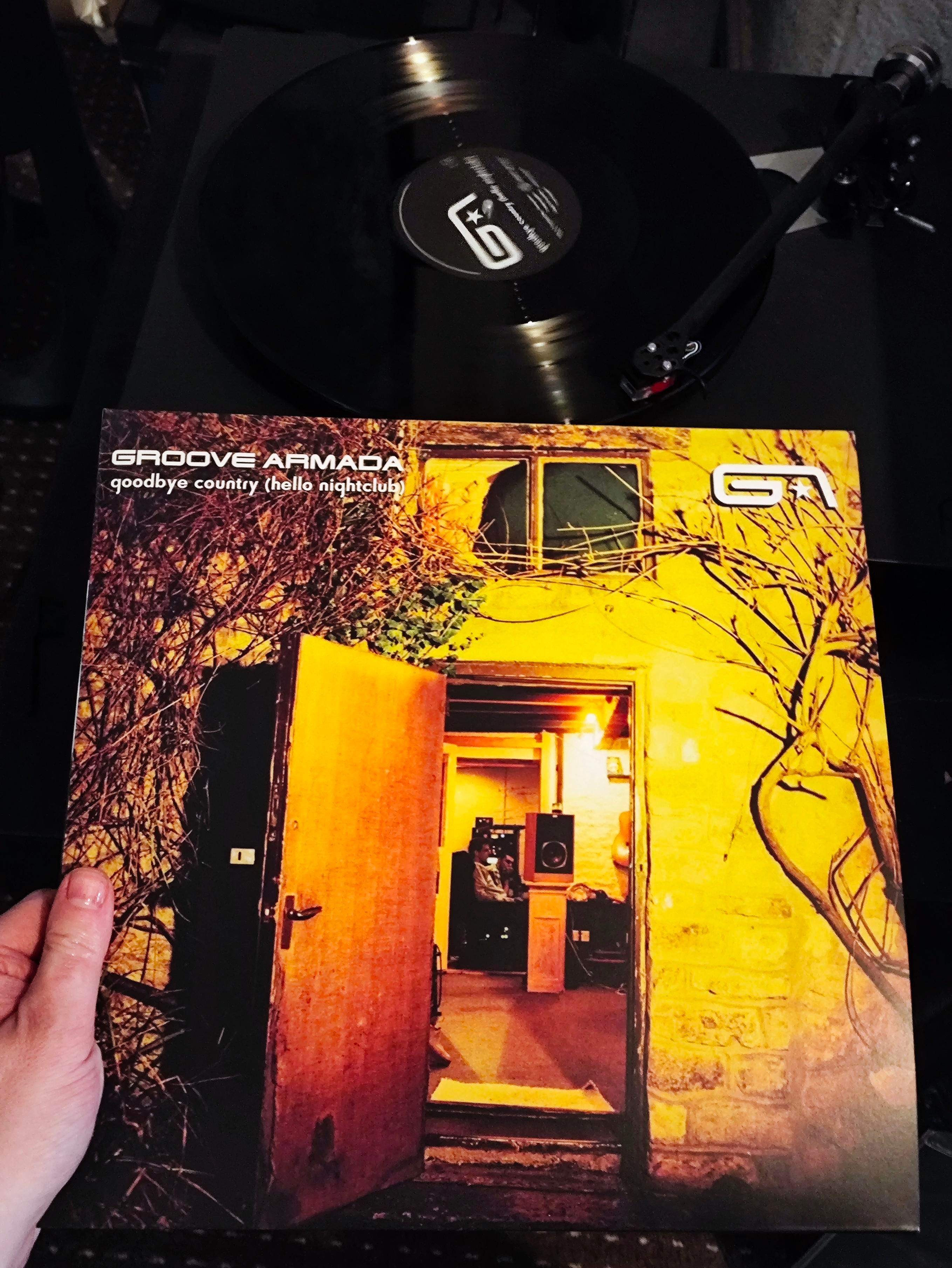A door on the outside of the house looking in to a studio where 2 people sit and a speaker is on furniture. Vinyl cover photo 