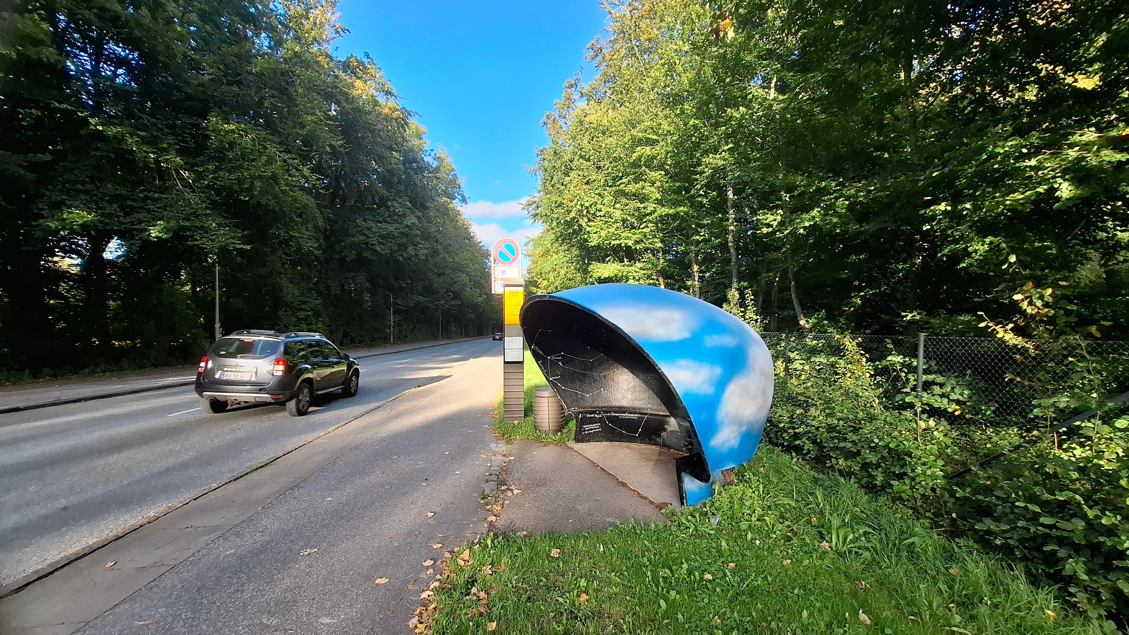 Seen from a side angle, so you can see the outside of the bus shelter with blue sky and white clouds painted on it, and night sky inside. Now follows same full description as on all photos in the toot, so skip if you’ve already heard it. Aarhus. On the road outside the Ole Rømer Observatory, named after another famous Dane, Carl Nielsen. A broad clear road in Kongelunden, the Royal Woods, with trees on either side. Looking straight at the bus shelter, one of the city’s iconic mushroom-shaped bus shelters designed in the 1960s by furniture designer Poul Cadovius. A low concrete mushroom-shaped shell with standing room and a little bench inside. The council commissioned artists to paint them with local motifs.  Artist Ekaterina Poliakova painted the night sky in this one. As you wait for the bus, you are surrounded on three sides and above by a black night sky with a map of the stars and constellations and galaxies, their names written beside them.  Outside, the shelter’s shell is painted like a day sky – light blue with fluffy white clouds. 