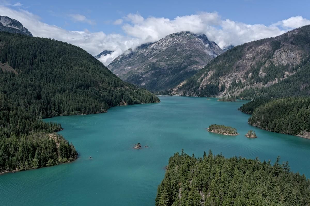 Color landscape photo of a lake in a mountain valley. The lake is turquoise in color due to minerals from melting glaciers. The lake is in the shape of an upside down "Y". At the head of the "Y" is a barely visible dam. The lake is wider where is disappears on the lower left and narrow where it disappears a bit below center on the right. A fairly flat piece of land sticks out from the lower right to about a quarter of the way up in the frame. Steep tree covered mountains are on the right and left. A tall cloud capped mountain is in the background under a blue sky.