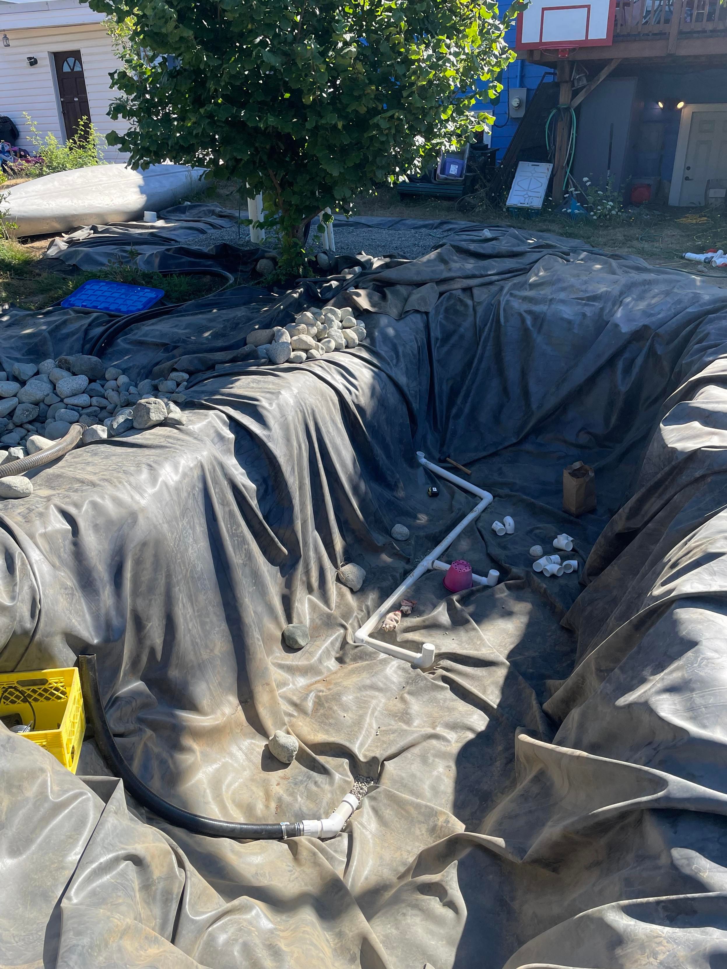A view into the black rubber lined hole shows  numbee of fittings lying in the bottom and some lengths of pipe fit together toward the back edge.