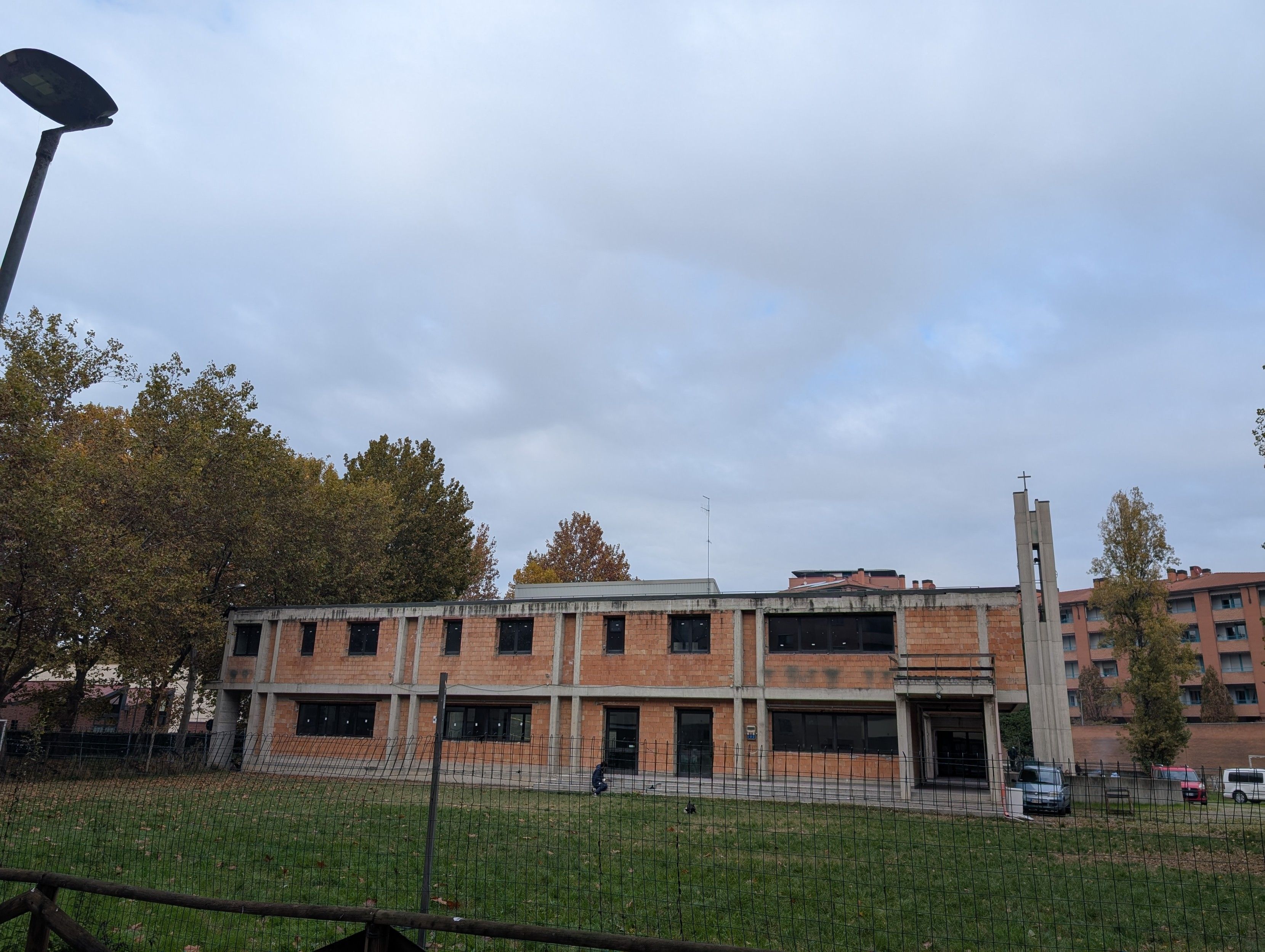 Foto di un edificio a due piani, con  la struttura di cemento a vista e le pareti di nudi mattoni rossi, sullo sfondo un campanile appartenente alla corrente traliccista.
