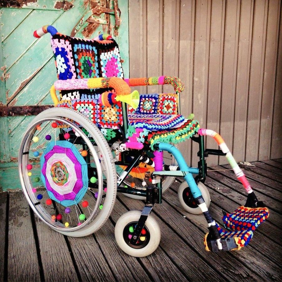 An old fashioned wheelchair with brightly coloured yarn and crochet covering the whole object in a way that joyfully transcends it