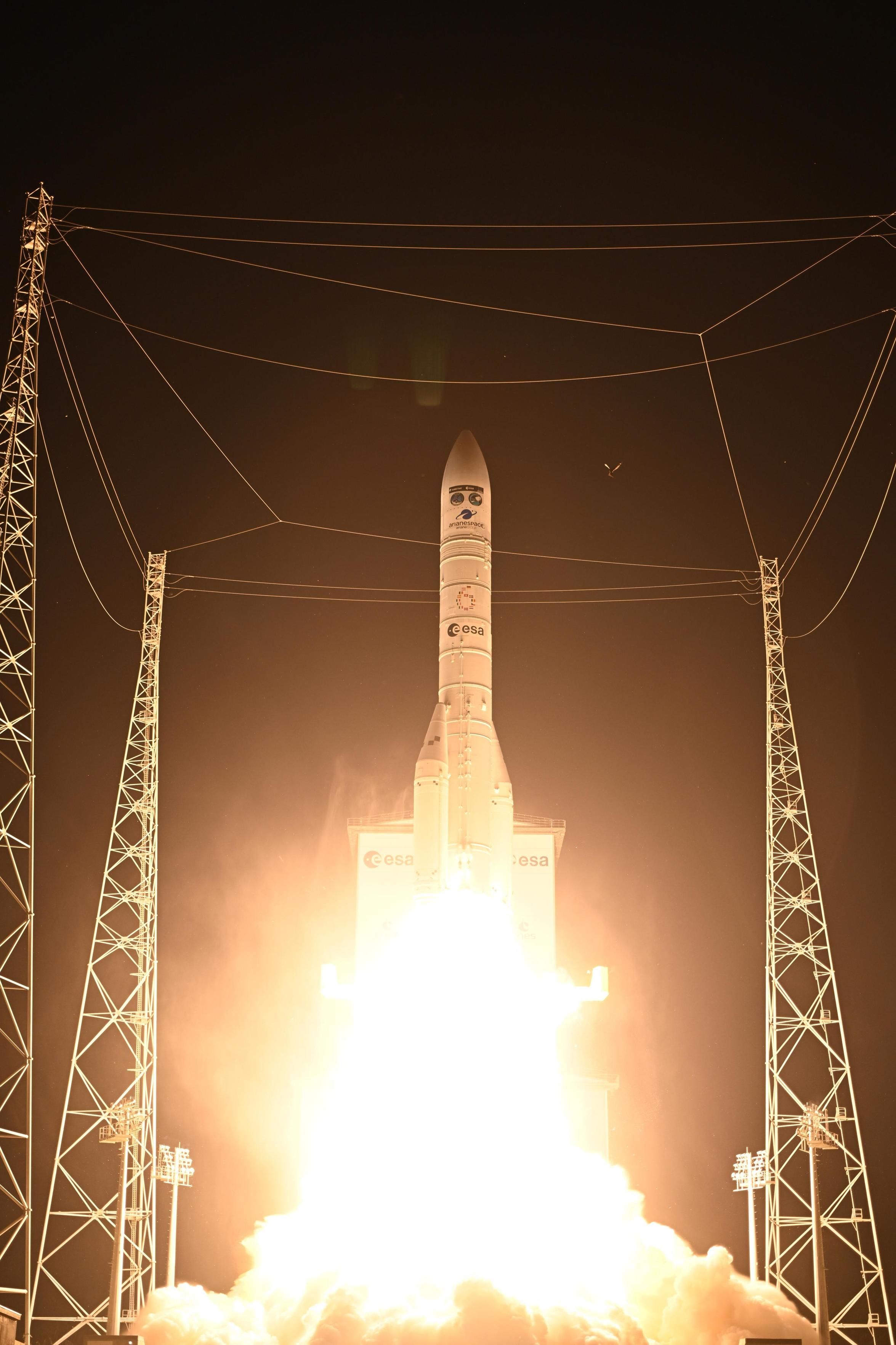 A Vega-C rocket launches at night from the European Space Agency’s facility, surrounded by tall support towers and cables, with bright flames and smoke erupting from its base as it lifts off.