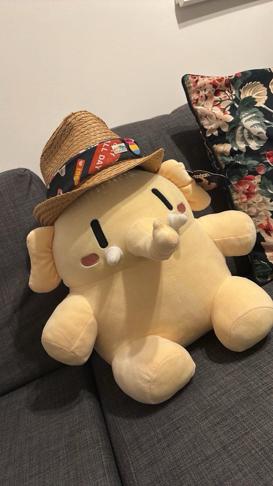 A large, plush mastodon toy wearing a straw hat sits on a gray couch, next to a decorative pillow with a floral pattern. The elephant has a soft, yellow body and cute facial features. The hat has two pins: „CSD-Salzburg“ and a Pangender Flag Pin