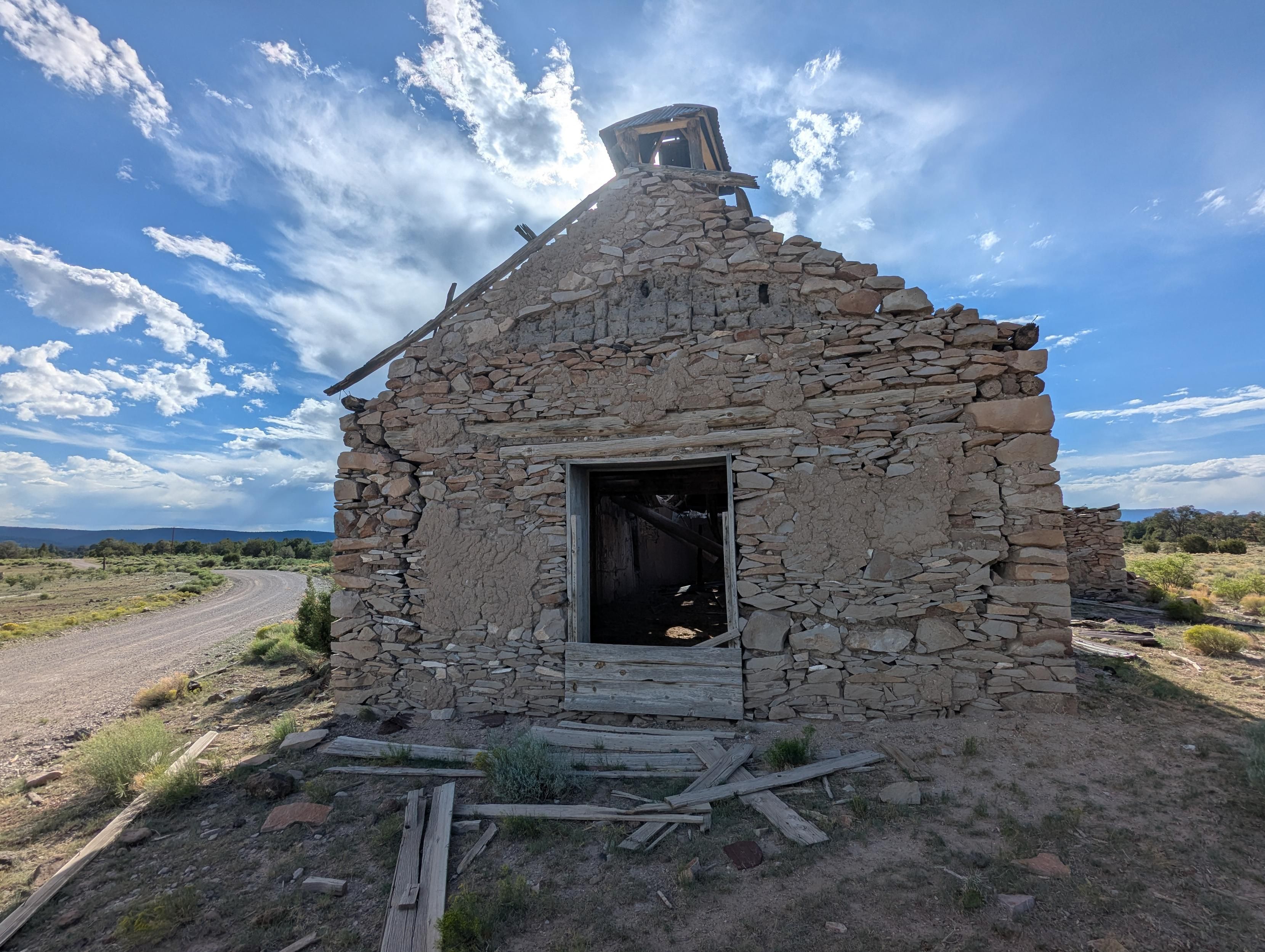 An old, falling-down adobe schoolhouse in rural New Mexico. The doors and windows are partially boarded up, but part of the bell tower on the roof remains. A gravel road passes on the left and curves into the distance. The sky is bright and partly cloudy, the desert is uncharacteristically green because of the monsoon rains. 