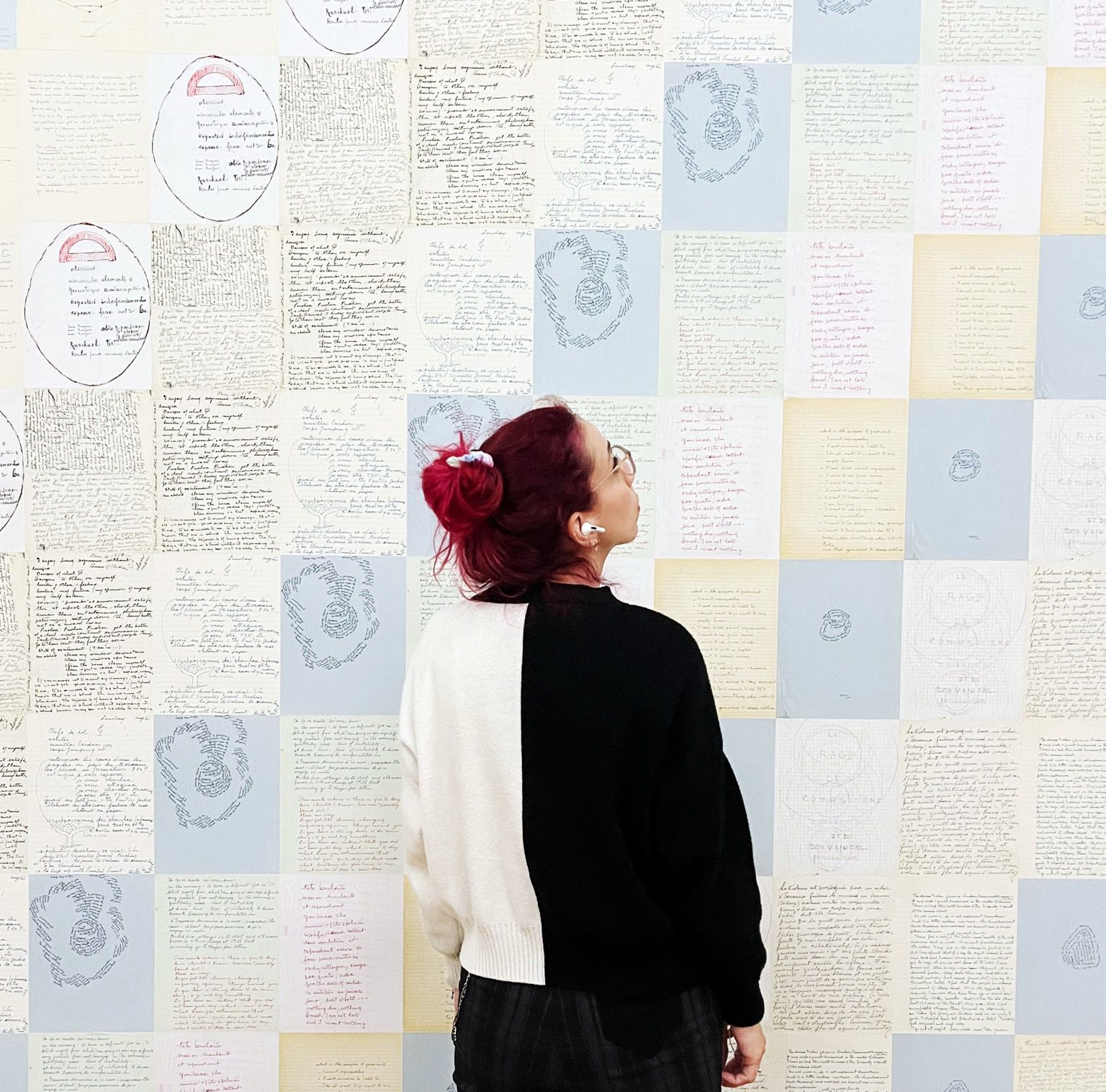 Je suis debout de dos face à un mur couvert de feuilles de papier manuscrites de différentes couleurs pastel. C’est une oeuvre de Louise Bourgeois. J’ai la tête tournée vers le haut, je regarde à droite. Je porte un chignon, des lunettes et un pull bicolore noir et blanc. L’atmosphère est contemplative et sereine.