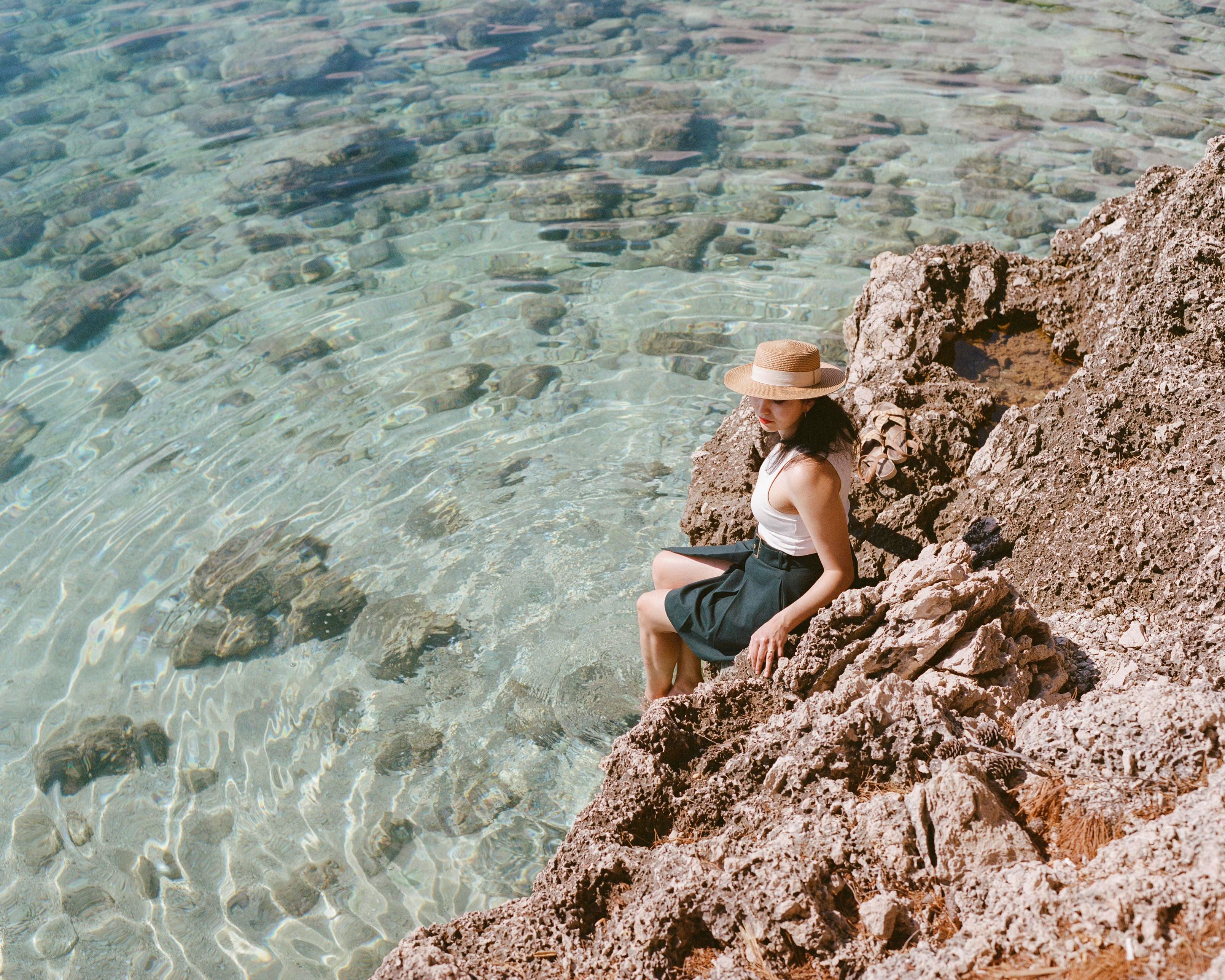 A woman dressed in a green skirt, white top and a straw hat is sitting on a rock formation on the coast with her legs in the water. The water is light blue and ripples are eminating from her legs through the whole frame.