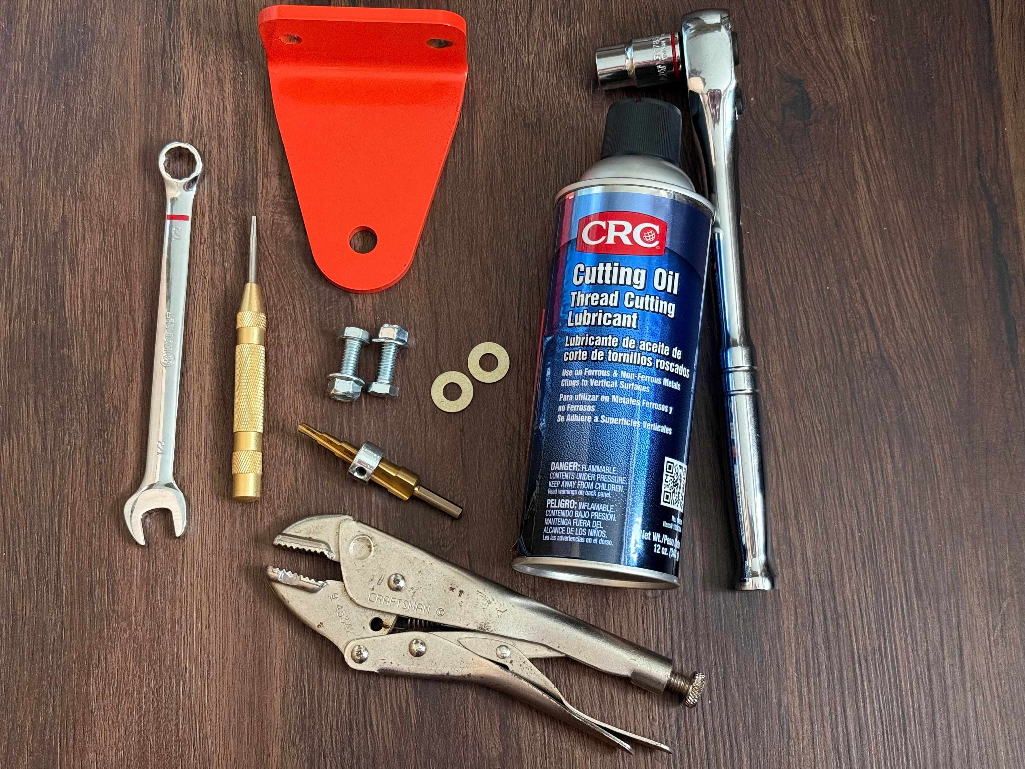A bright orange hitch plate with pre-drilled 5/16 inch holes a half inch wrench to hold the nut a half inch socket wrench to hold the bolt two washers two nuts and bolts, cutting oil spray can multi step drillbit with a stop above 5/16. And my grandpa’s very old craftsman vice grips 