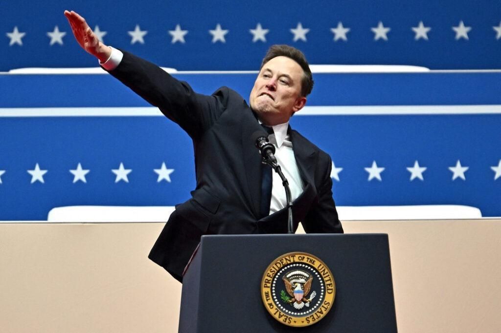 White male (Elon Musk) in suit and tie, behind a podium with the Presidential seal giving a Nazi salute. 