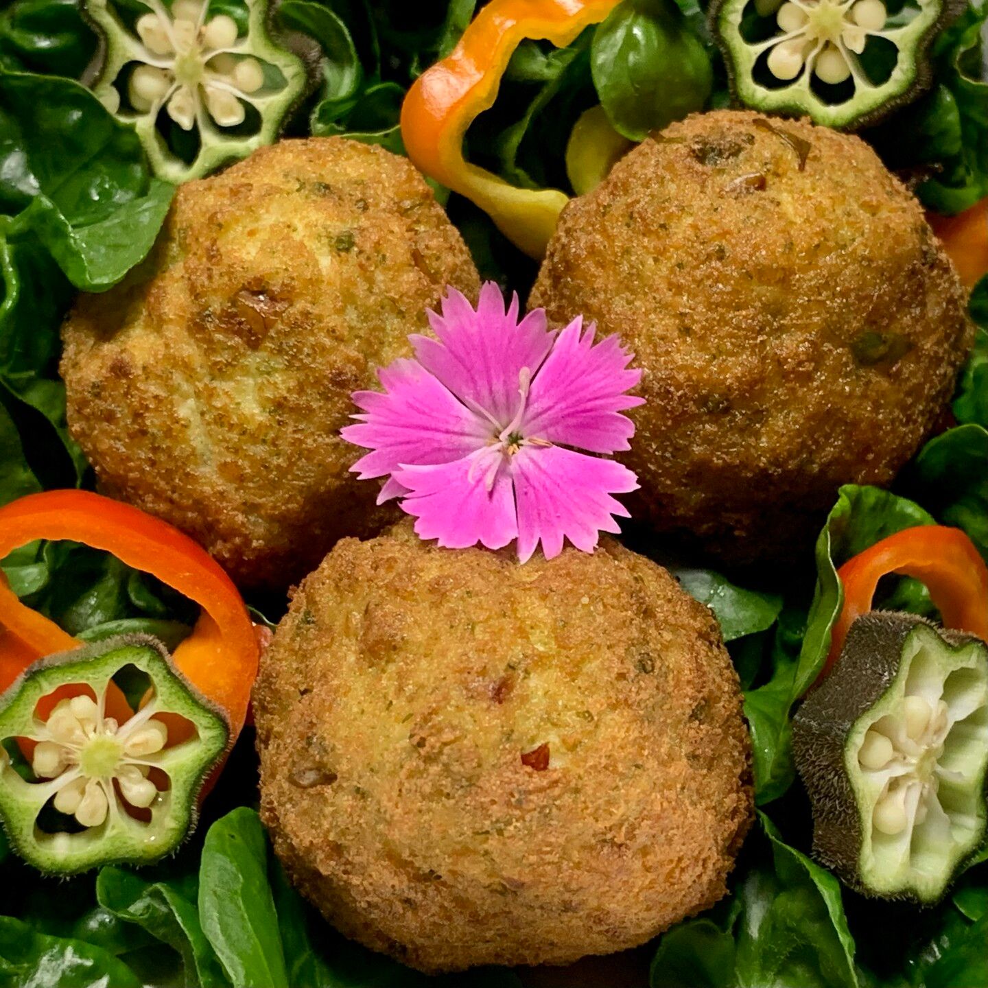 On a bed of curly green sissoo leaves, sliced green okra and small sliced red and orange madhatter peppers are three green fried tofu balls. They are browned but beneath the dried exterior is a subtle green hue. In the middle of the fried tofu is a bright pink dianthus flower. The ragged edges of its petals contrast with all the round elements in the image. 