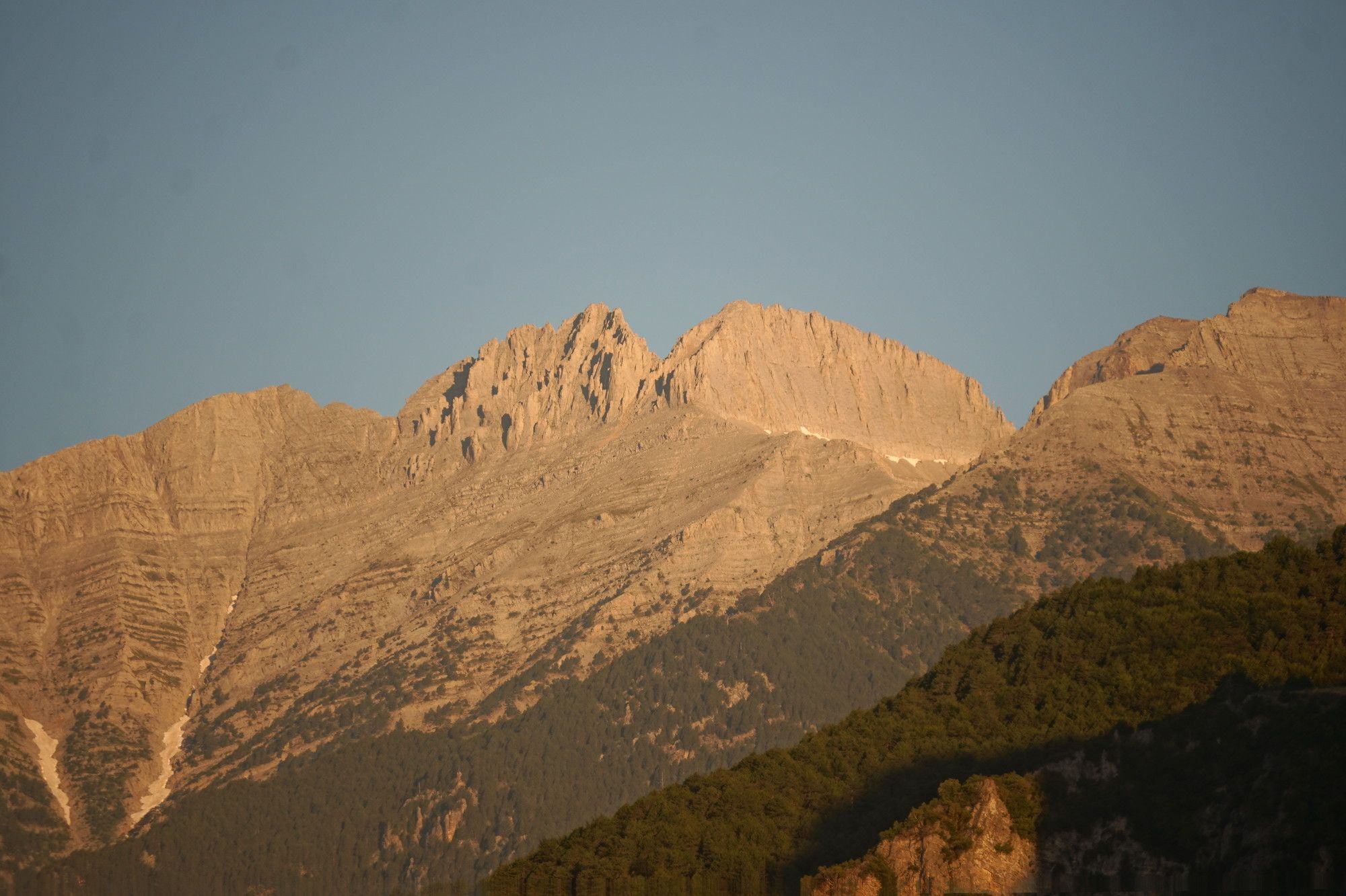 The two biggest peaks of Mount Olympus, Mytikas and Stefani, are kind of beige-coloured with a blue sky behind them. There is sort of a V notch between the rocky peaks. At the base you have some trees. 
