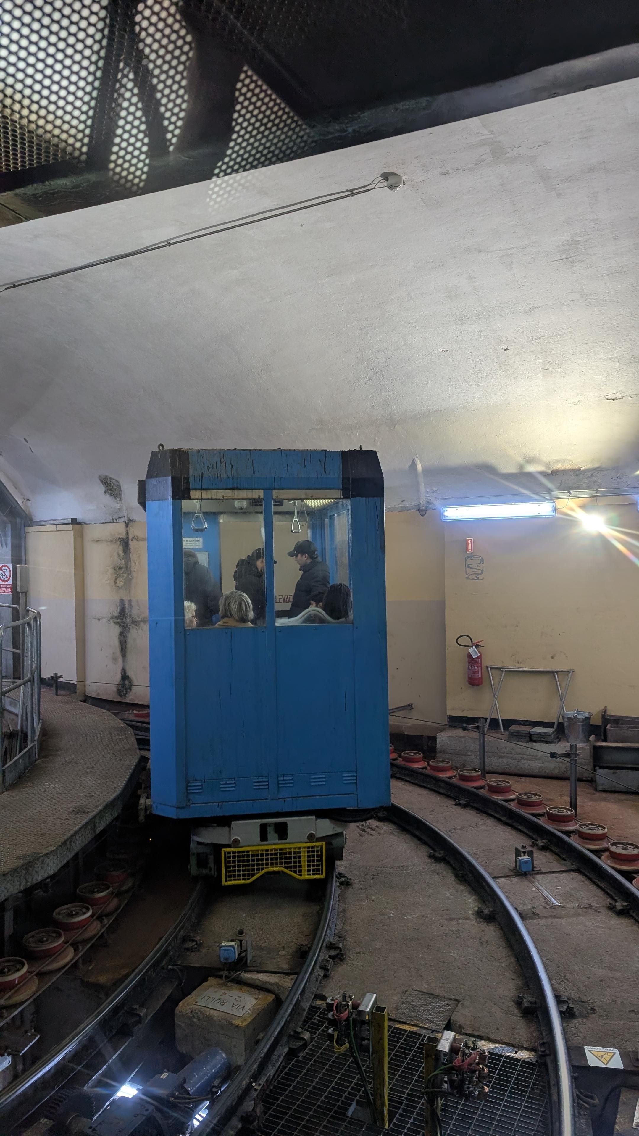 A blue, cubic cabin on a bogie, two tracks merging in a narrow radius in an underground room, red spools along the outer sides of the track