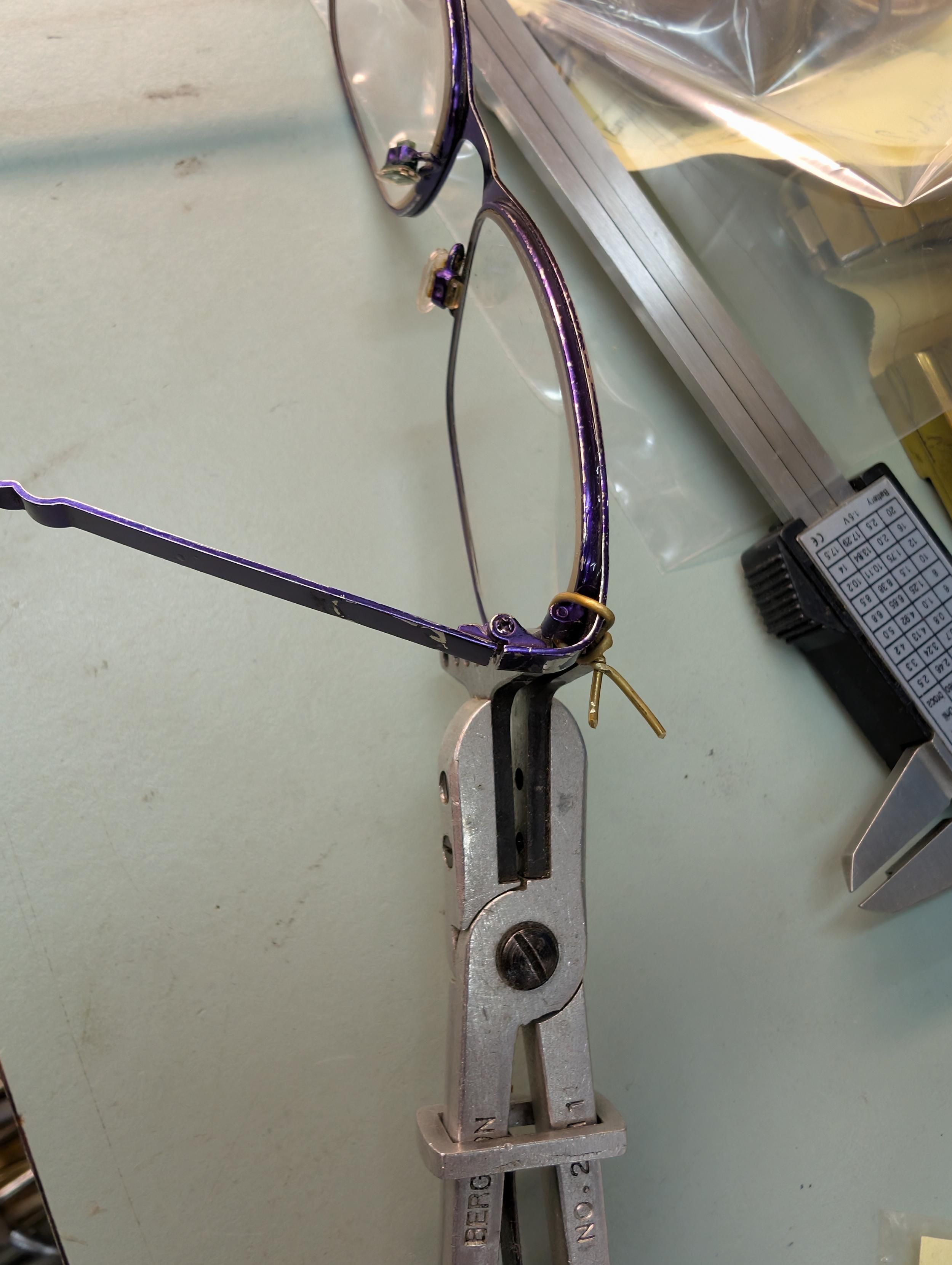 The side of a purple pair of glasses with a bit of brass wire, twisted to help hole the frames together and slide pliers to hold the frame together too
