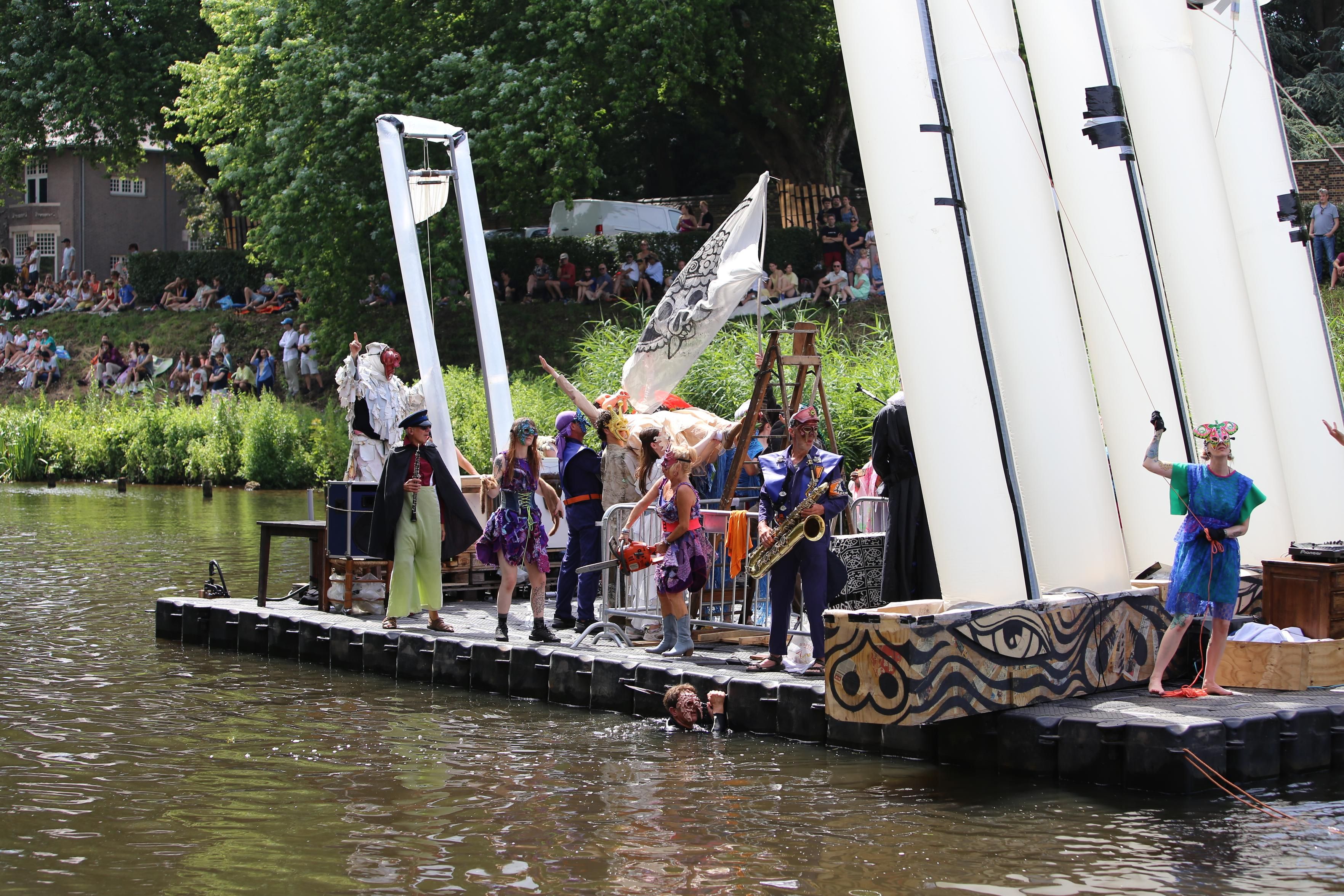 Barge with an inflatable guillotine and people wearing odd outfits. Some have instruments and one has a chain saw. A person in a pig mask is crowd surfing.  Swimmers are towing it through the canal. 