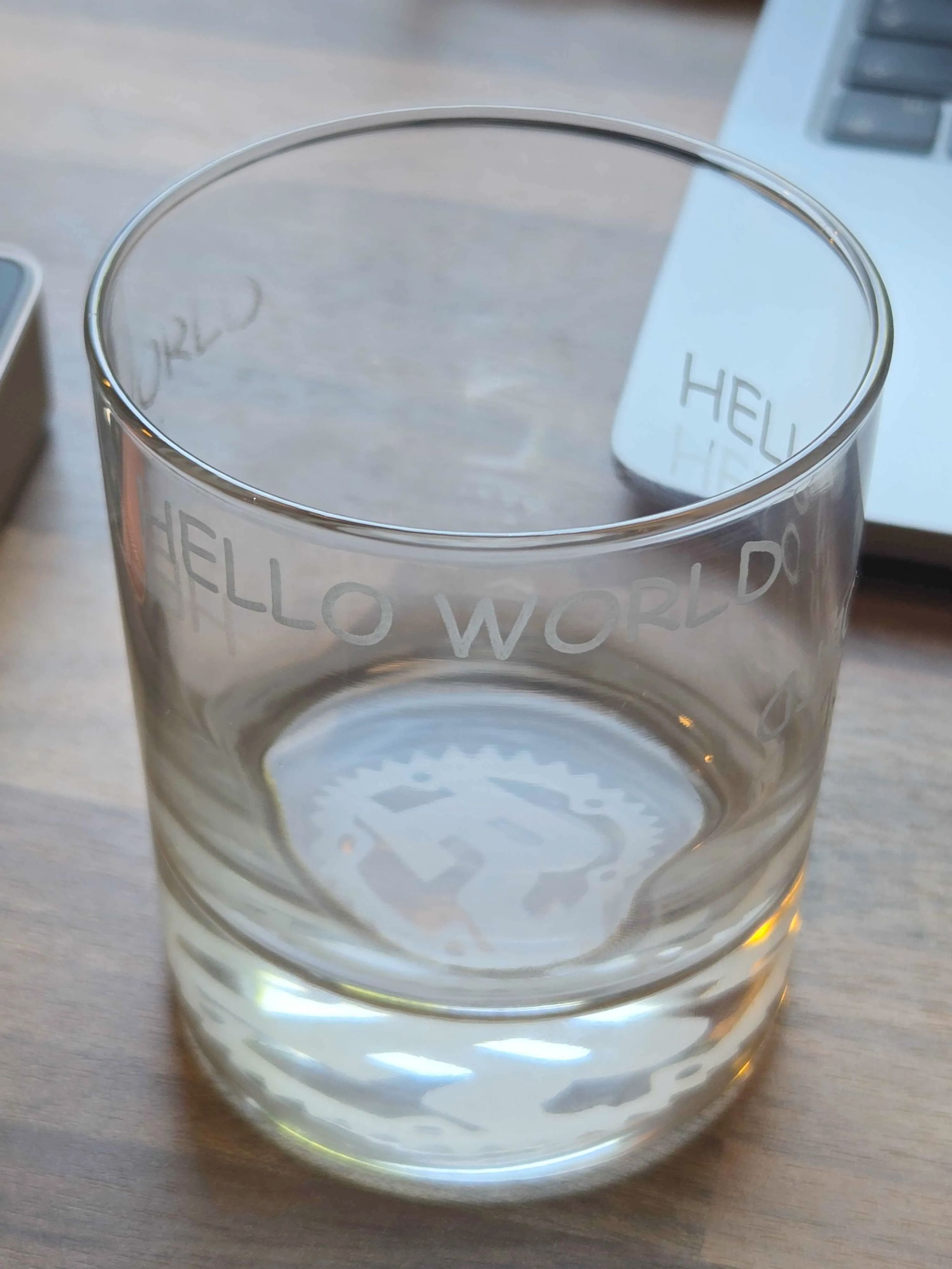 A whiskey glass with "HELLO WORLD" engraved on it in Comic Sans, in several places. The bottom of the glass has the Rust logo engraved in it.