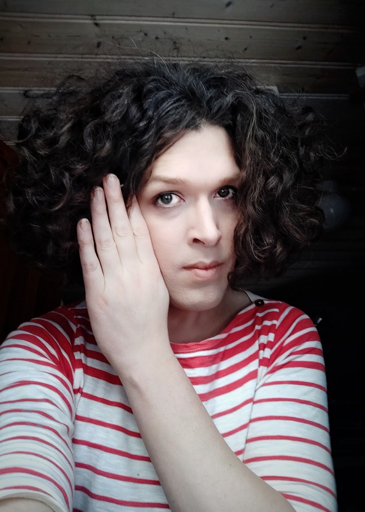 Selfie, moving my hair out of my face with my left hand, looking into your eyes, my mouth is closed, my curly dark brown hair is fluffy and hangs almost to my shoulders, my top is striped white and red.