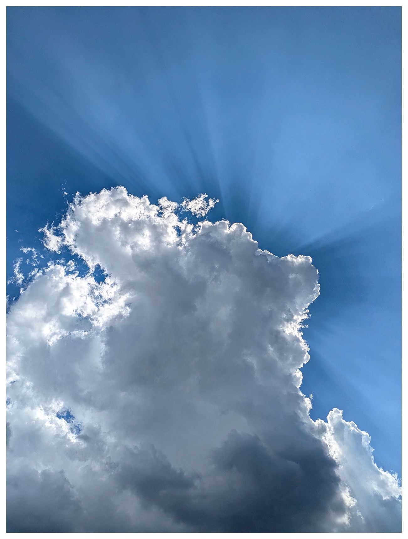 ai-assisted alt text

A large, bright white cloud dominates the center of a blue sky. Sunbeams radiate dramatically from behind the cloud, creating striking rays of light. The cloud’s edges are illuminated, and the sky is clear except for a few wisps of cloud.