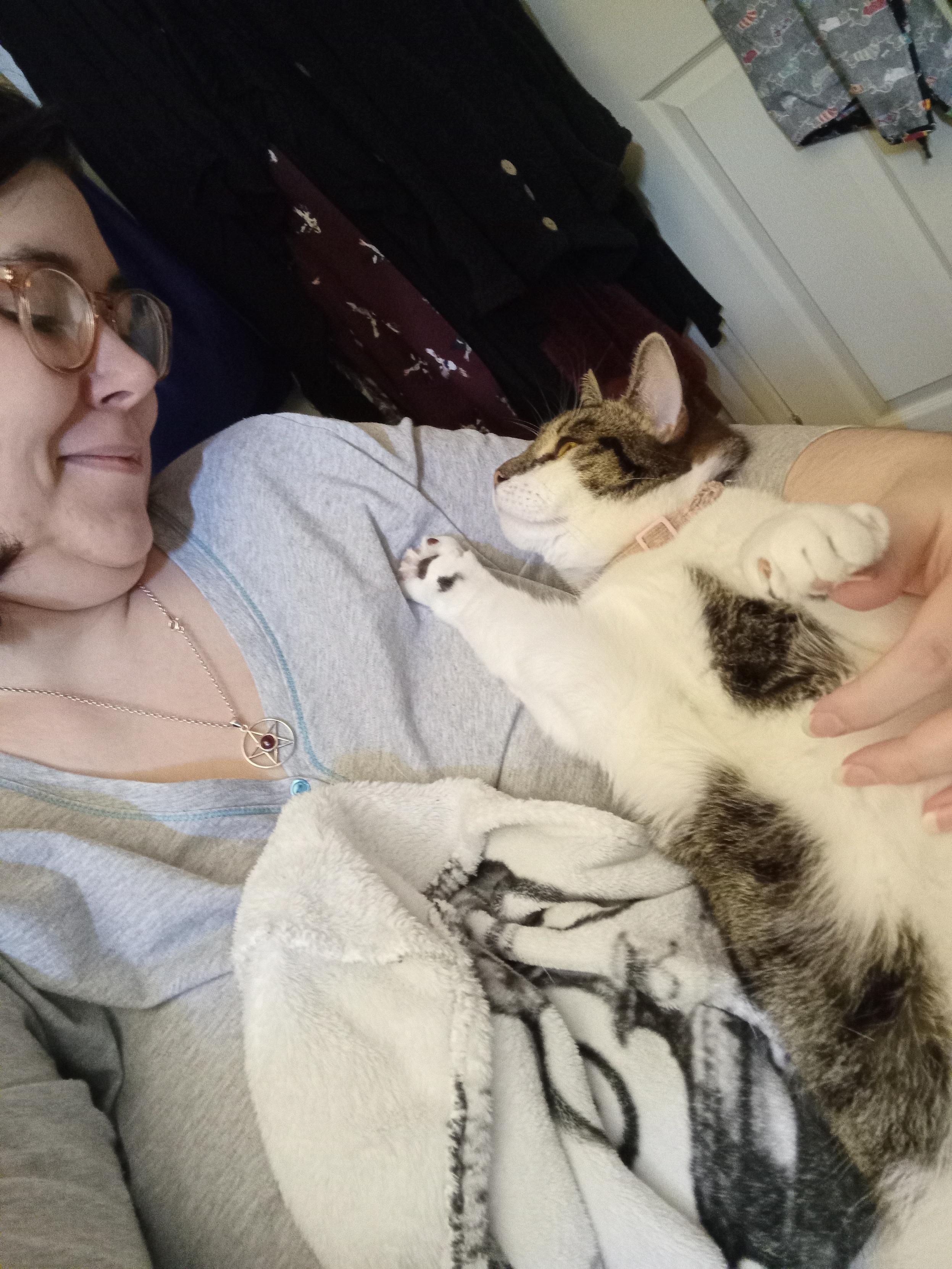 Me, a white enby with glasses, smiling as she peers up at me from the crook of my arm whilst making biscuits in the air.