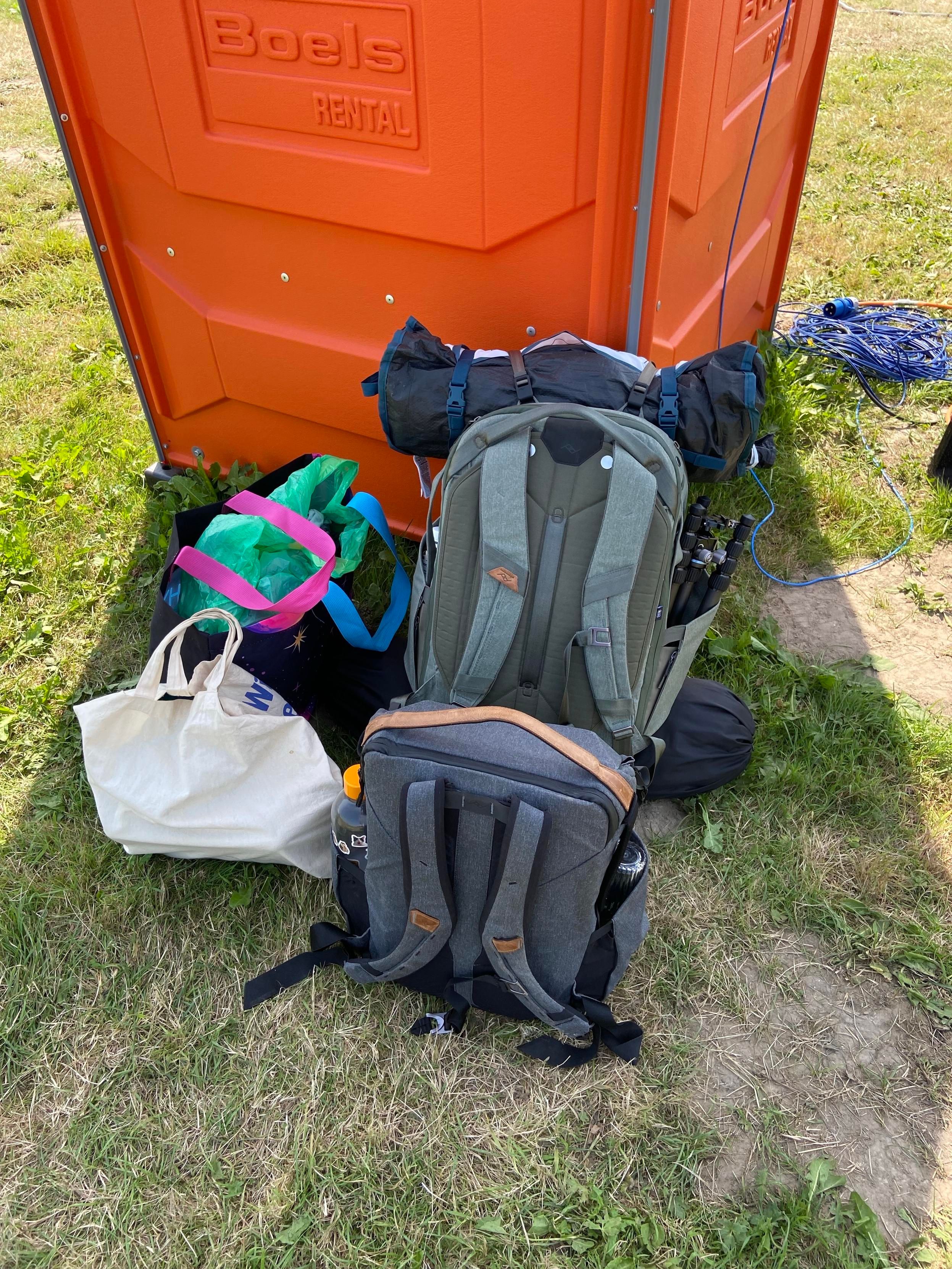 A photo of my luggage: a jute bag, a WHY bag (from the merch desk), a large backpack with things strapped to its top and bottom, and a medium backpack.
They are resting in the shade behind a Datenklo.