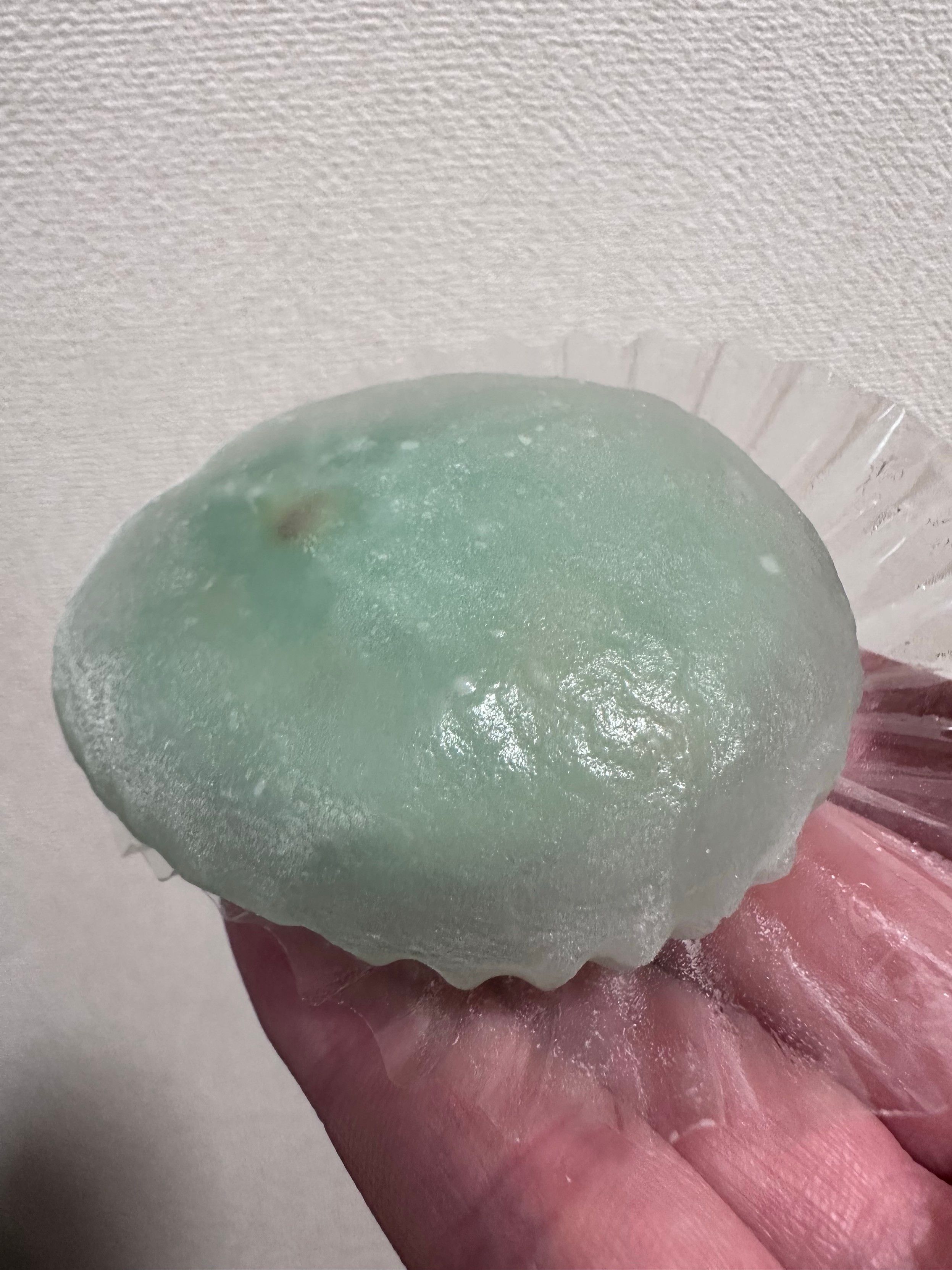 A hand is holding a large, oval-shaped dessert with a translucent green exterior. The dessert appears to be a type of mochi, served on a paper liner, and is set against a light-colored background.