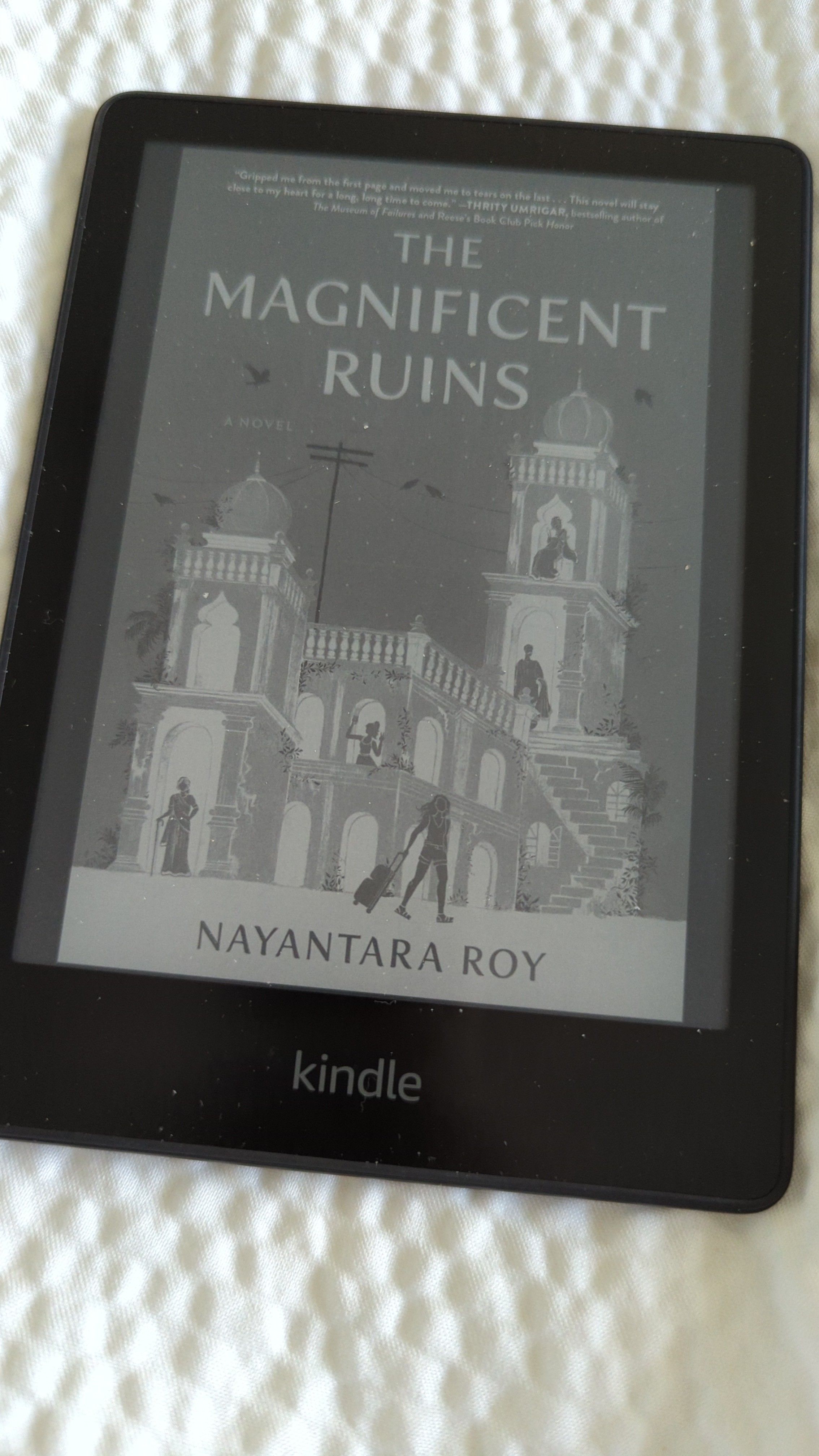 A black kindle showing the cover for The Magnificent Ruins by Nayantara Roy which depicts an Indian palace with two turrets and lots of doors/windows with silhouetted people sitting or standing in 4 openings and a silhouetted woman in the foreground with a rolling suitcase.