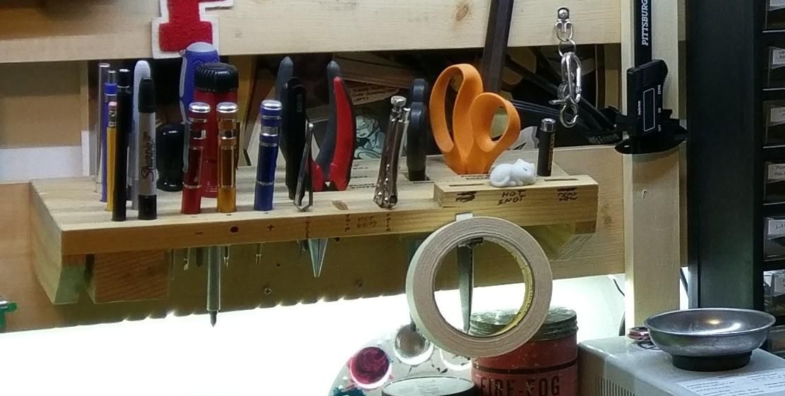 A piece of wood mounted to another piece of wood at a slight angle, with several holes cut or drilled into it and an array of pens, pencils, screwdrivers, scissors, and pliers resting in it. A roll of masking tap hangs from the front and a small white toy cat sleeps peacefully over the handwritten words "hot snot" on the end.