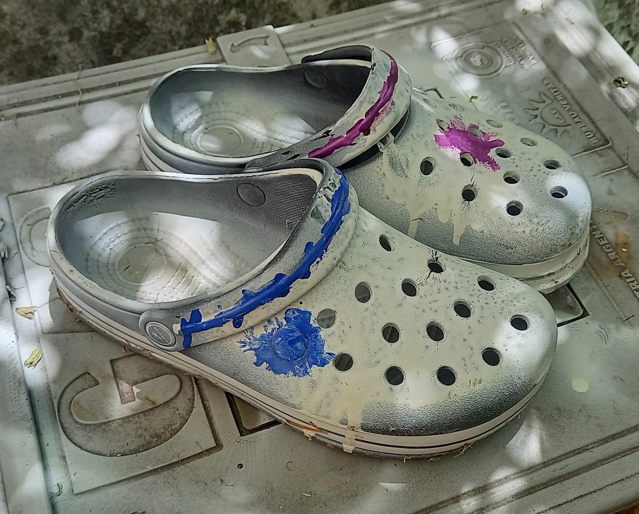 Crocs spray-painted white from above and to the right. Nearest has a blue strip along its heel strap and an irregular spot in the same color; farthest is the same but in purple. They're on the same surface where they were sprayed, a plastic door for a gas meter box.
