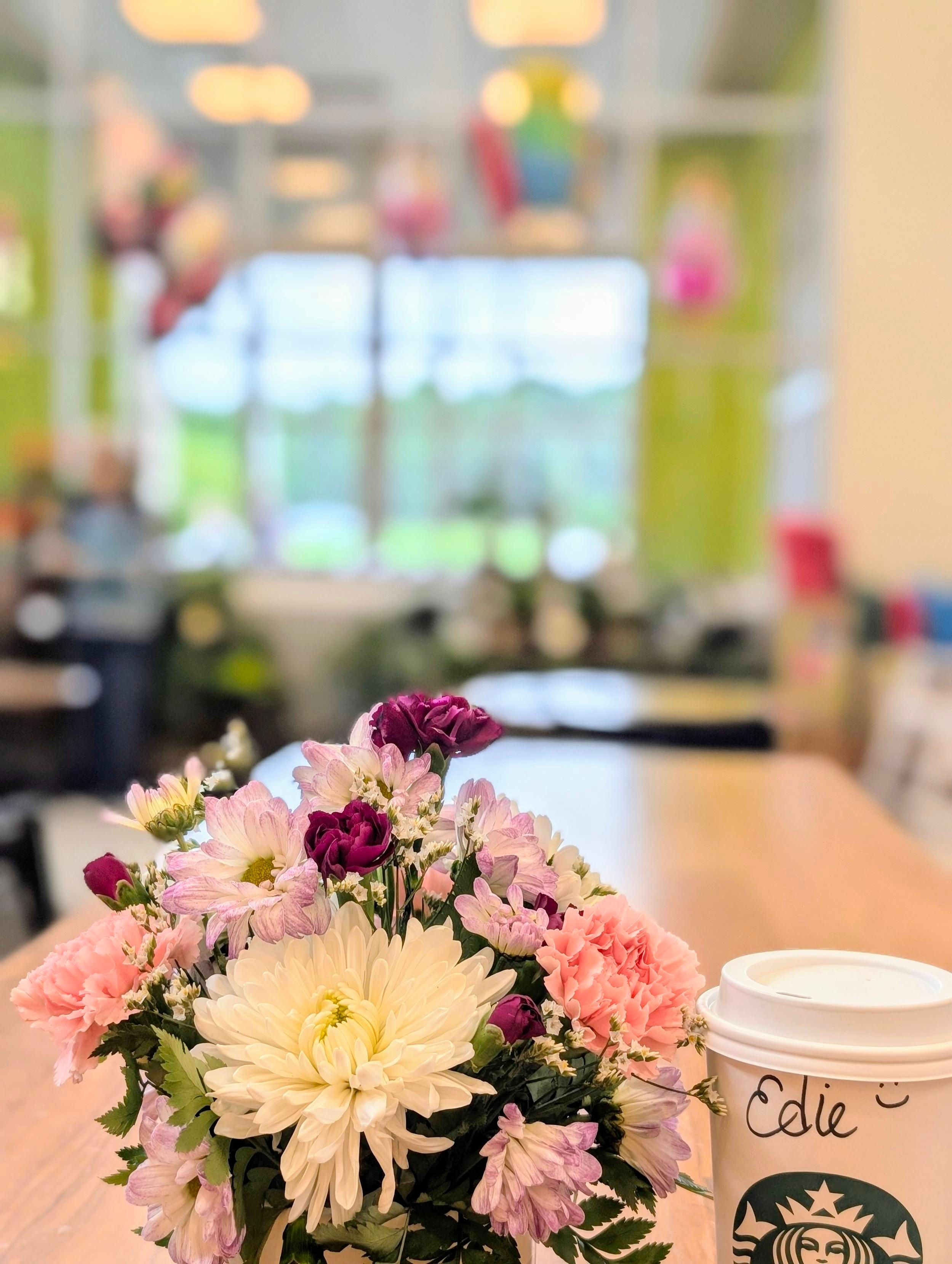 Small arrangement of mums and carnations with bits of fern sits next to a carryout coffee cup on a long wooden table out of focus in the background. large windows with balloons and flowers in front