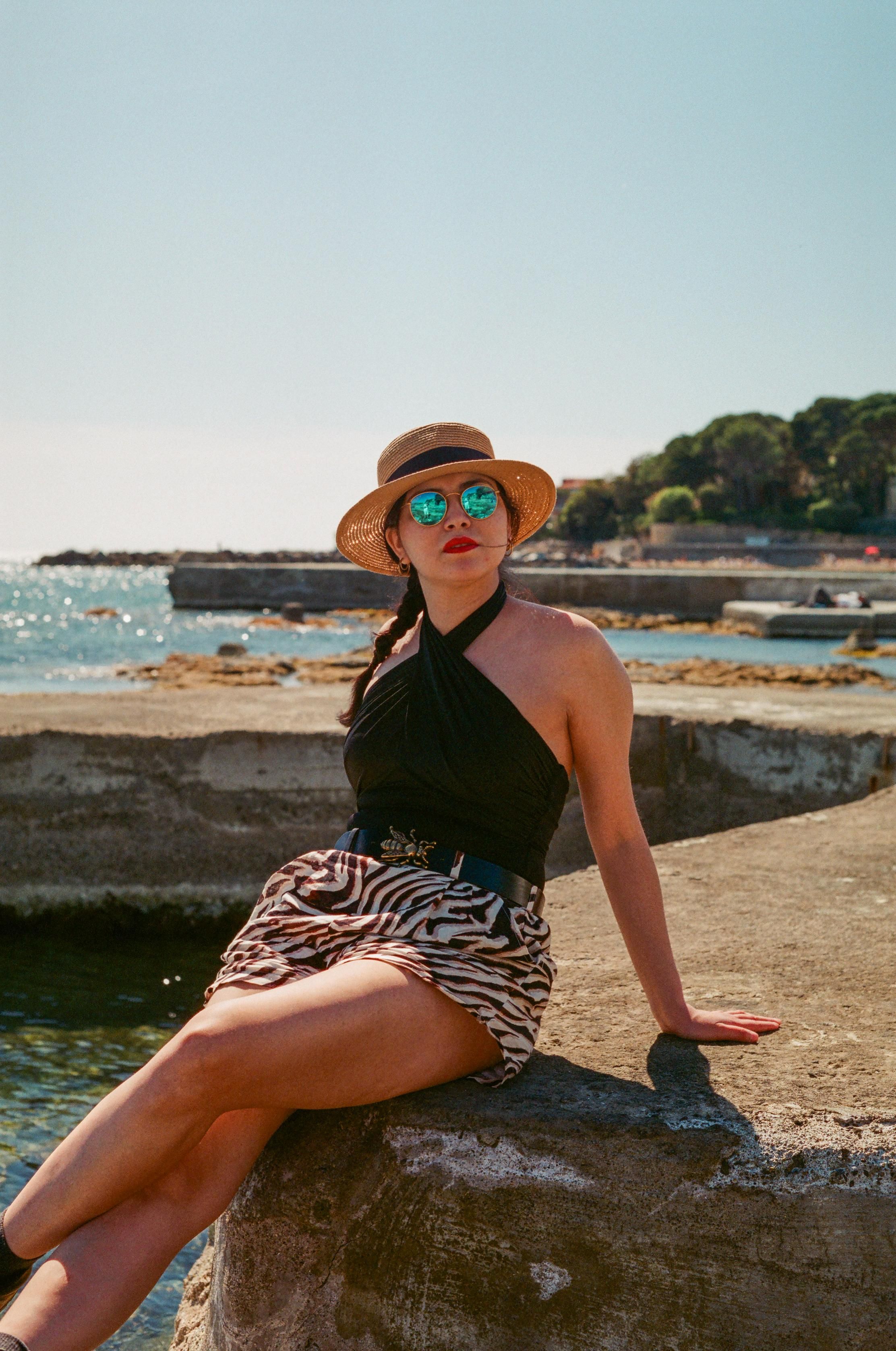 A beautiful woman wearing a straw hat, a black cross-halter neck top, animal print shorts, a belt with a metal bee, and reflective sunglasses, sitting legs crossed on a concrete pier in front of a Mediterranean coastline.