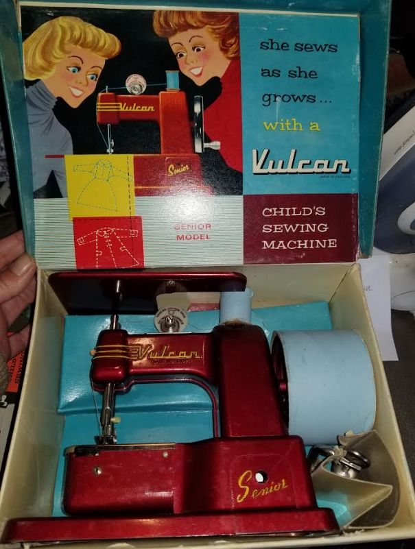 A vintage toy sewing machine, inside the packaging box. The lid is being held open. Inside the lid is a very 1950s style drawing of two girls looking at a sewing machine. Text reads She sews as she grows with a Vulcan child's sewing machine.

The machine itself is a rich shiny red, and is lying flat in the box. It is shaped like an older model machine, with the word Vulcan in yellow on the top bit, and the word senior in yellow on the base. 