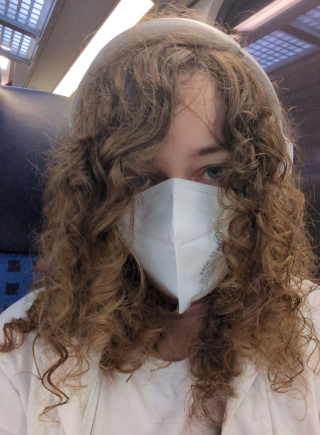 alina wearing a white mask, lab coat and headphones to supress the noises of the train she's in. her hair is especially pretty here