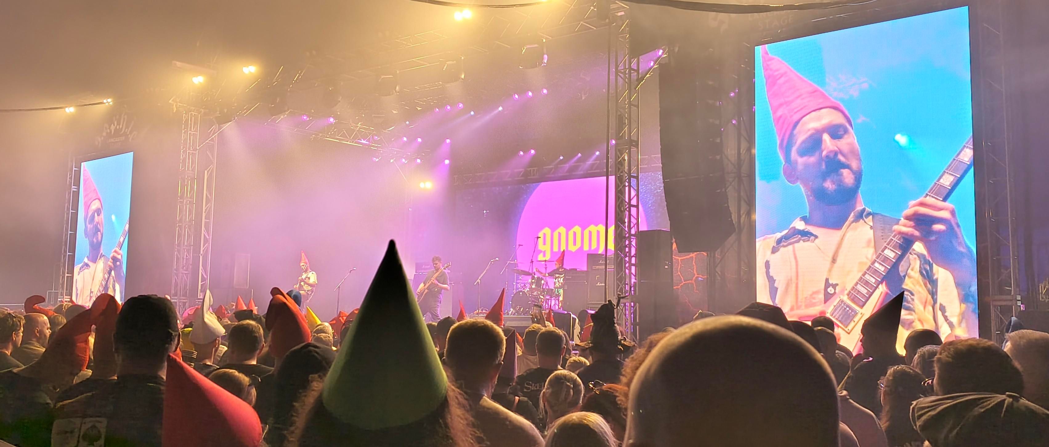Gnome the band sporting red gnome hats on a large screen next to the main stage. With a crowd alsk in gnome hats