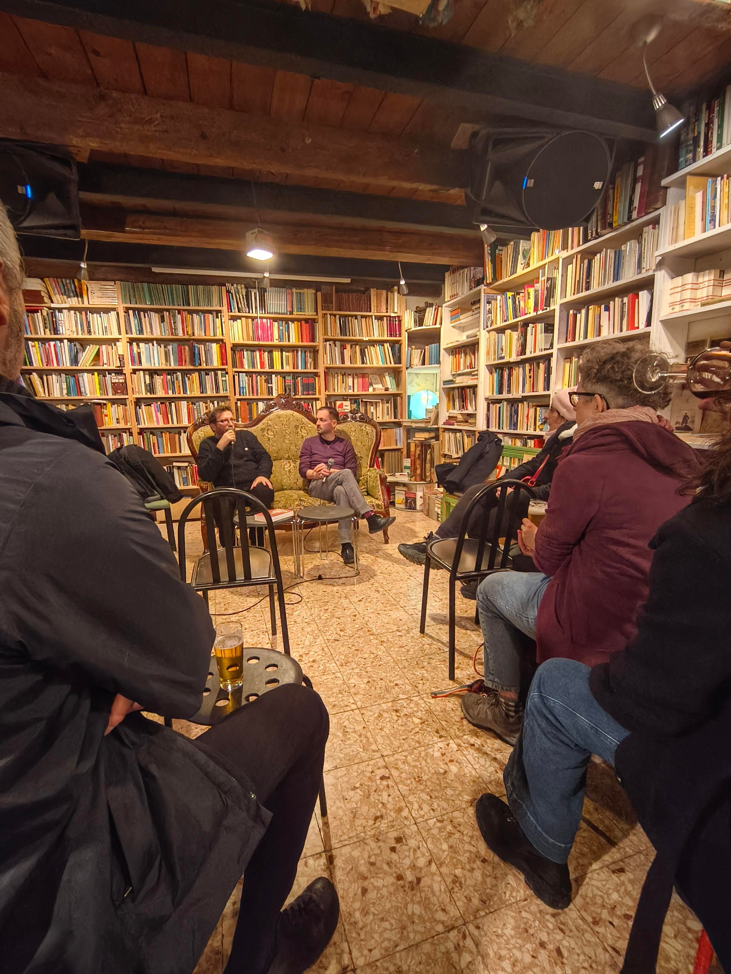Un divano con i due relatori, all'interno di una delle sale della libreria. Le pareti sono ricolme di libri. In primo piano parte del pubblico seduto di spalle.