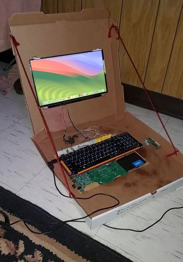 picture of a laptop that has been torn apart and has its internals placed inside of a pizza box. its all wired up and opened and powered on, displaying the macos sonoma desktop on the lcd display