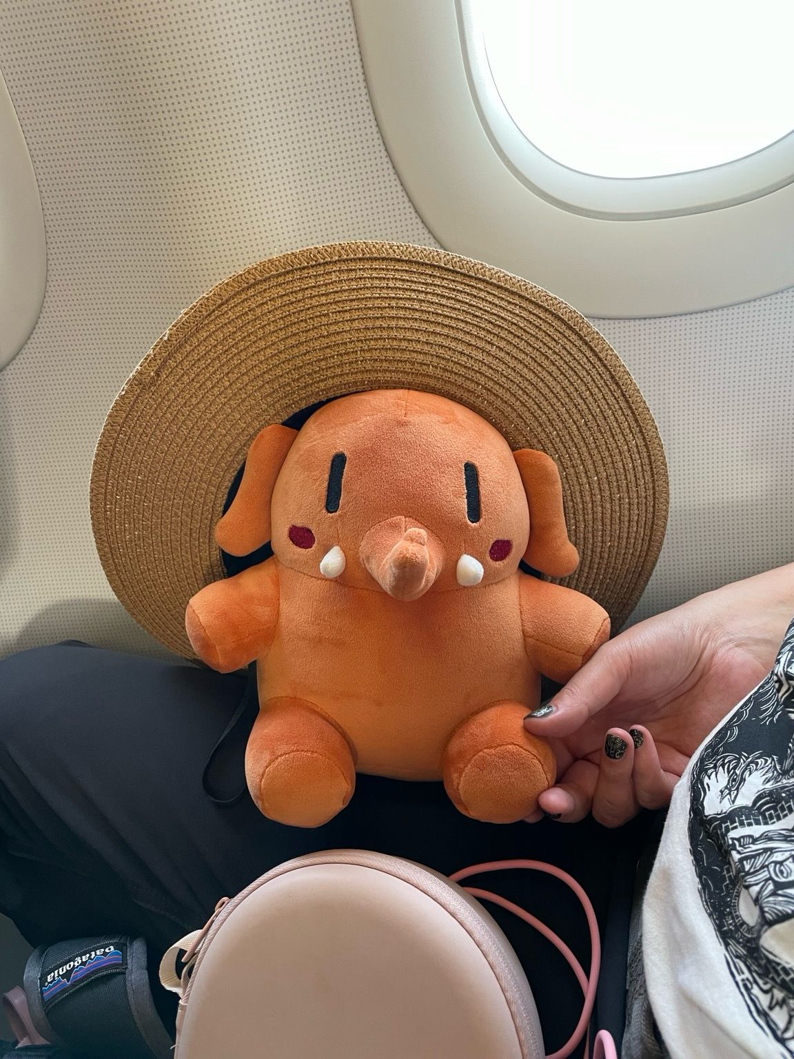 An apricot-colored stuffed Mastodon toy wearing a straw hat that is much larger than him inside a plane.