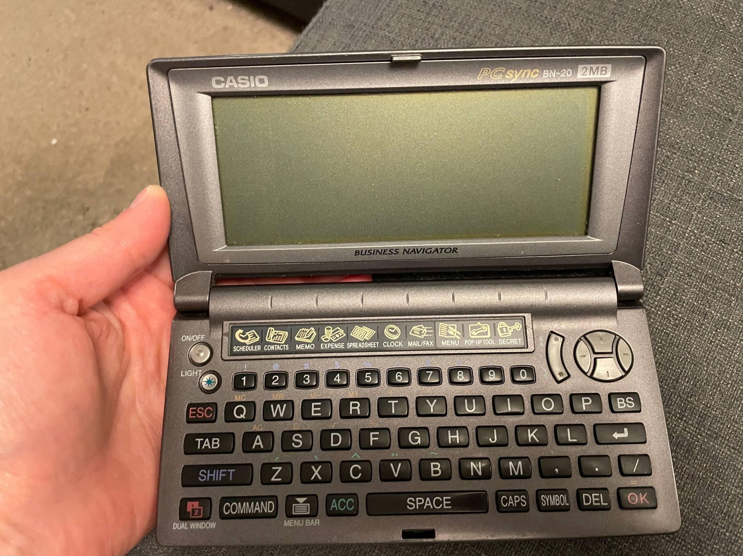 A photo of a palm-top computer, on top of a human palm. It is a business organiser device with a QWERTY keyboard and a large LCD screen. The label says "Casio PC Sync BN-20 Business Navigator 2MB"