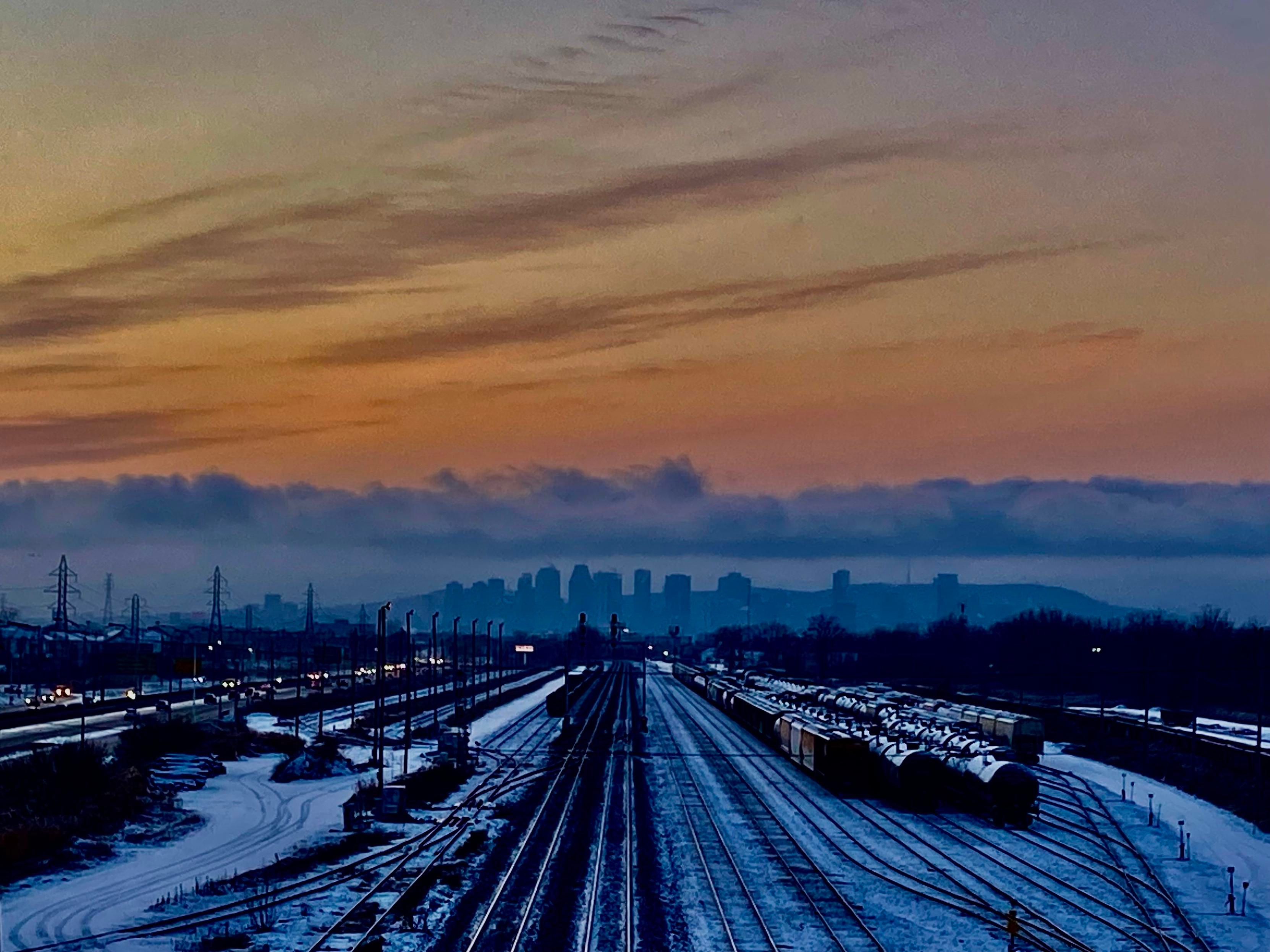 Railroad tracks in the foreground lead from a train yard to the Montreal skyline on the horizon. The island appears to be against a backdrop of white mist, under a low cover of clouds. Above the clouds, the sky is orange and yellow with purple streaks of clouds. 