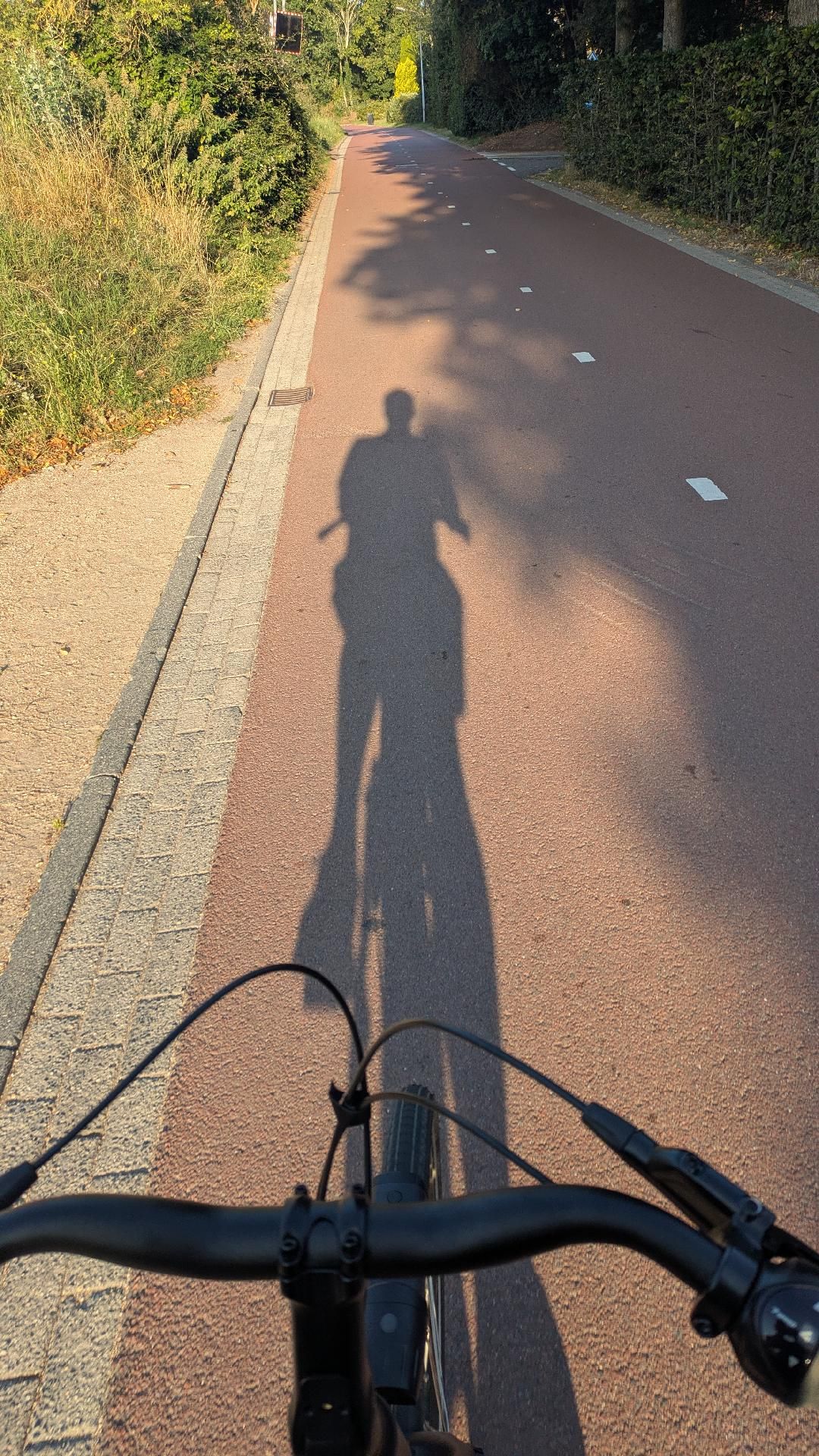Schatten von mir auf einer roten Fahrradstraße. Im Vordergrund ist ein Fahrradlenker zu sehen.
